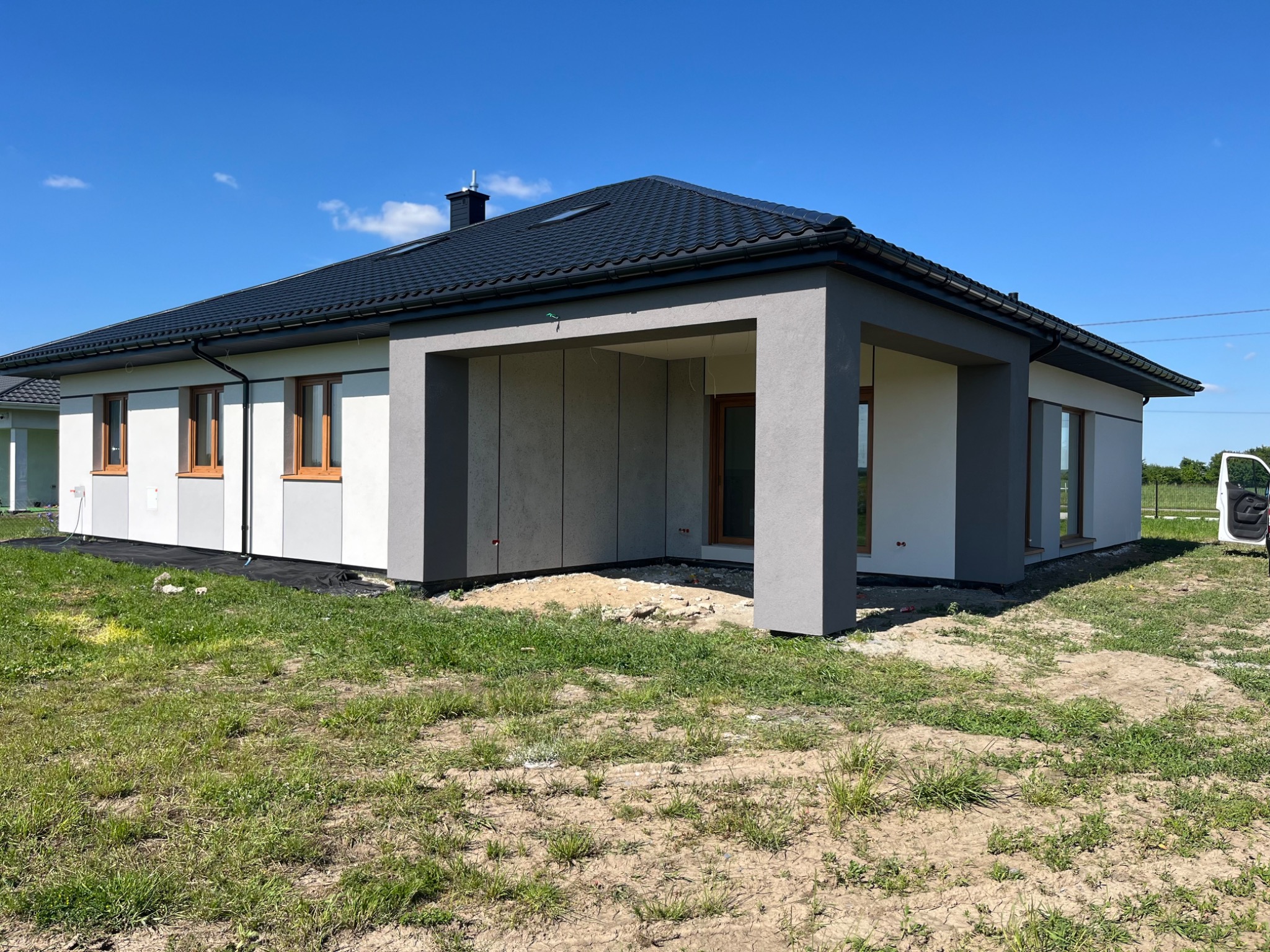 Nowoczesny dom jednorodzinny z grafitowym dachem i jasną elewacją, częściowo wykończony, z widocznymi oknami z drewnianą stolarką i fragmentem trawnika.