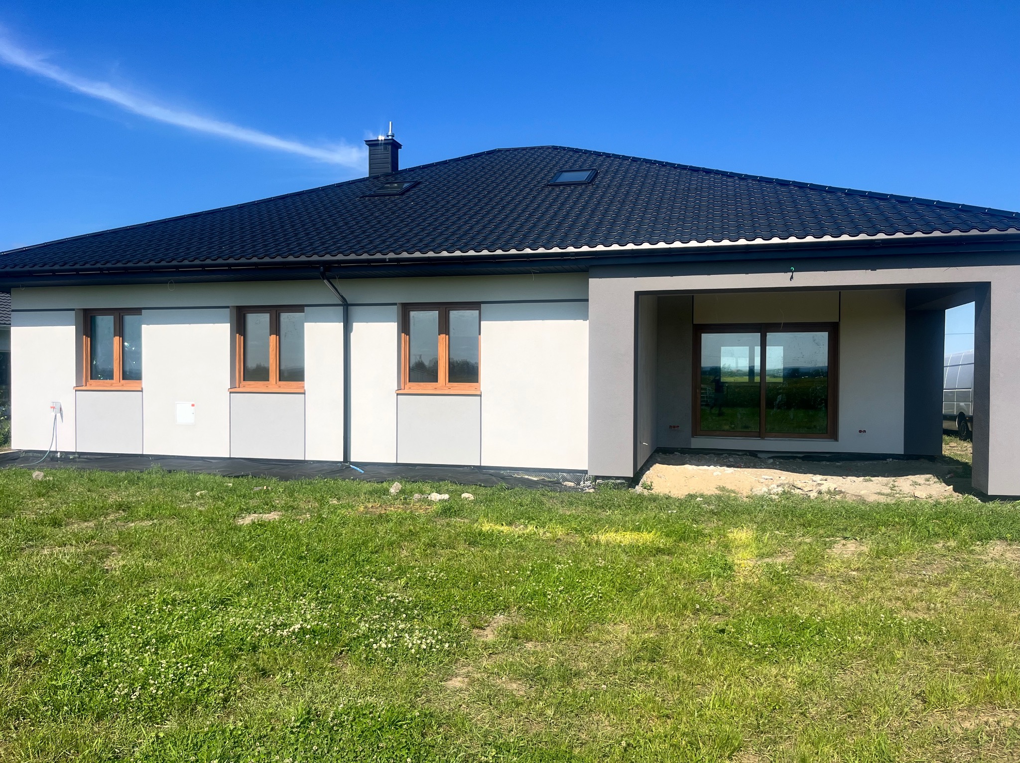 Nowoczesny dom jednorodzinny z ciemnym dachem i jasną elewacją. Widoczne drewniane okna i fragment ogrodu z zieloną trawą. Błękitne niebo w tle.