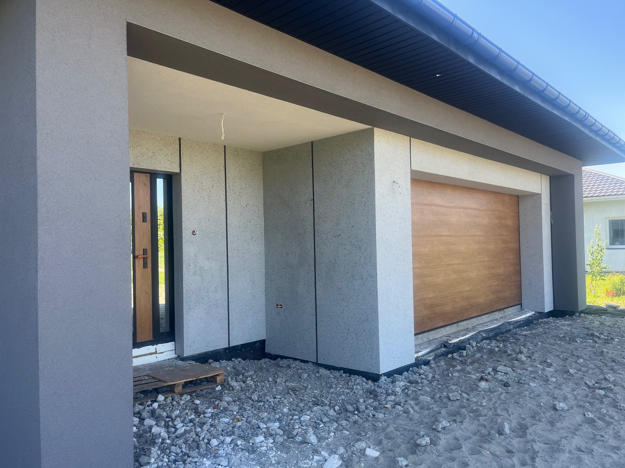Szara elewacja domu z drewnianą bramą garażową i drzwiami wejściowymi. Widoczne wykończenia i faktura tynku. Teren wokół domu w trakcie prac wykończeniowych.