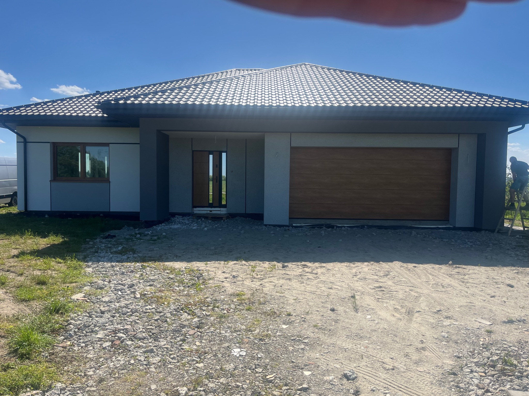 Nowoczesny dom jednorodzinny z szarą elewacją i drewnianą bramą garażową w słoneczny dzień. Widoczny fragment drabiny z pracownikiem.