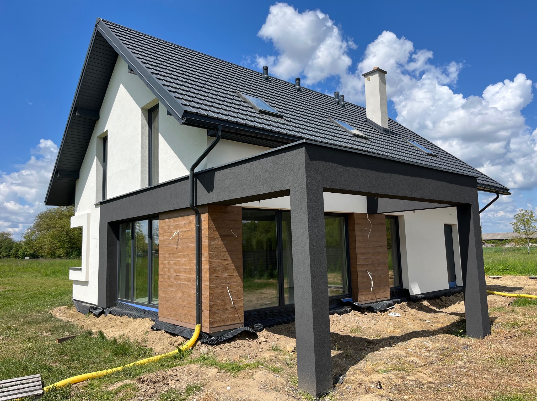 Nowoczesny dom jednorodzinny z ciemną dachówką i drewnianą elewacją, częściowo otynkowany na biało, z widoczną instalacją odprowadzającą wodę.