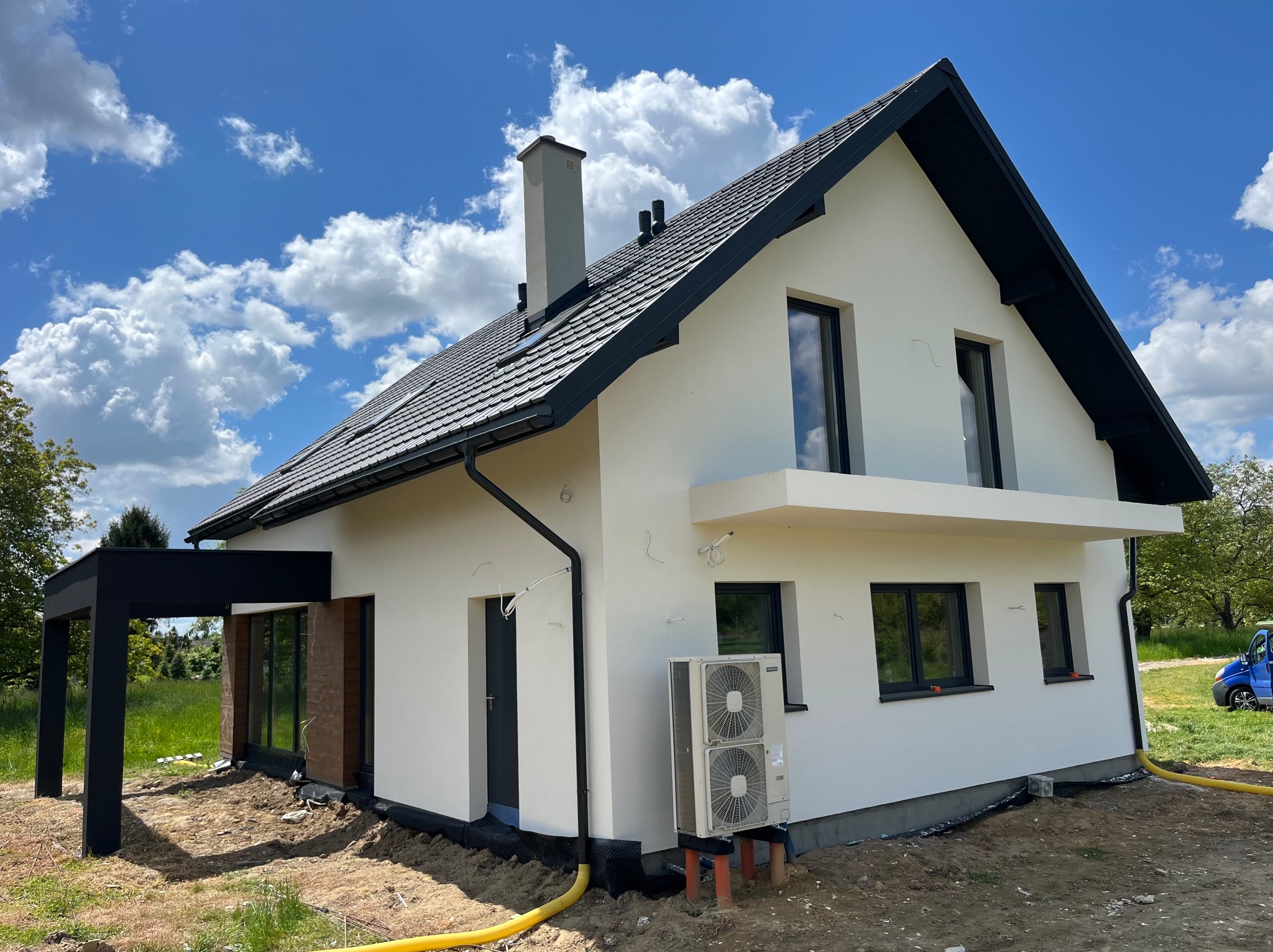 Nowoczesny dom jednorodzinny z białą elewacją, czarnym dachem i stolarką okienną, w otoczeniu zieleni i błękitnego nieba z chmurami.