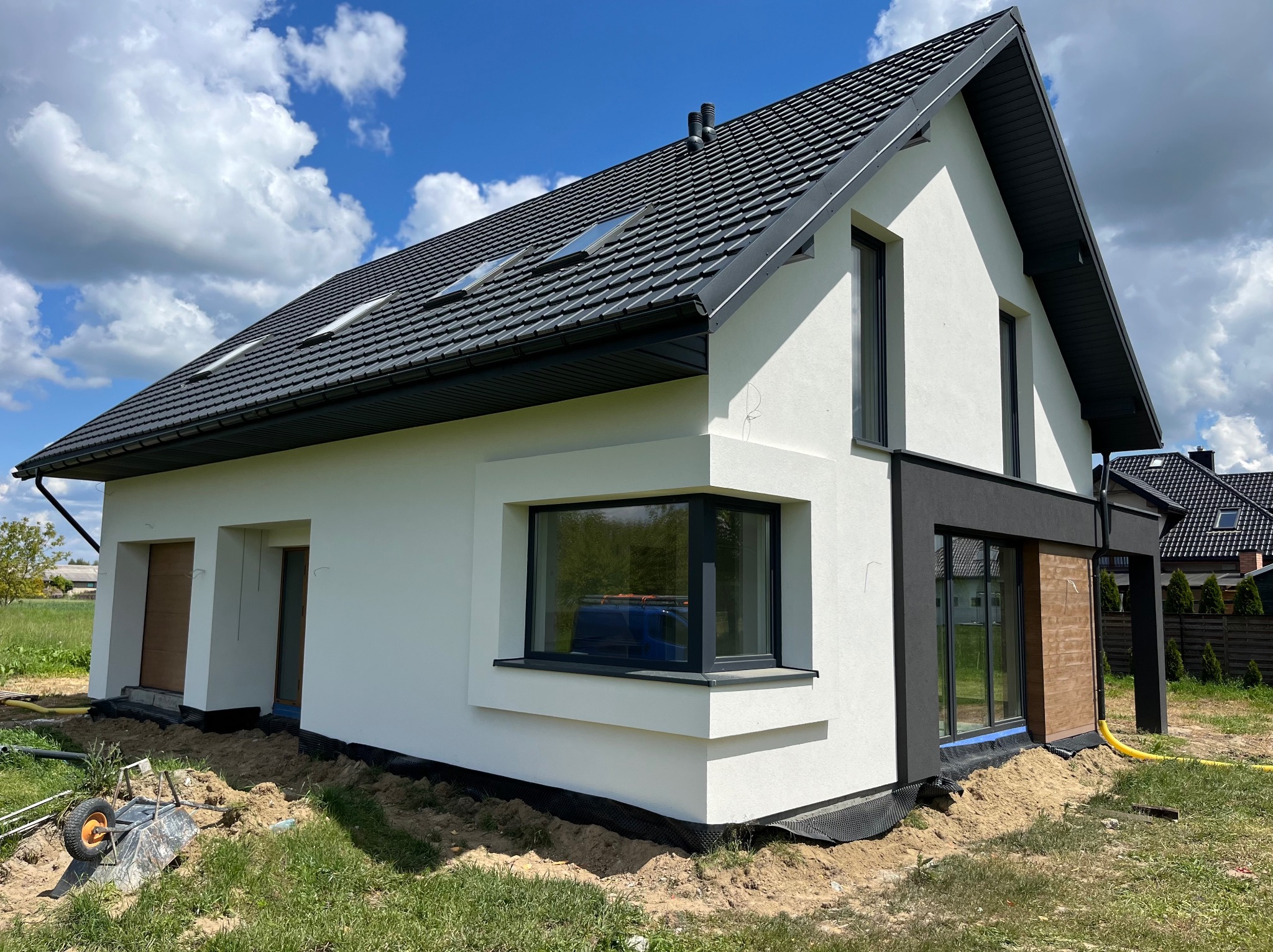 Nowoczesny dom jednorodzinny z białą elewacją i grafitowym dachem na tle błękitnego nieba. Widoczny wózek budowlany sugeruje prace wykończeniowe.