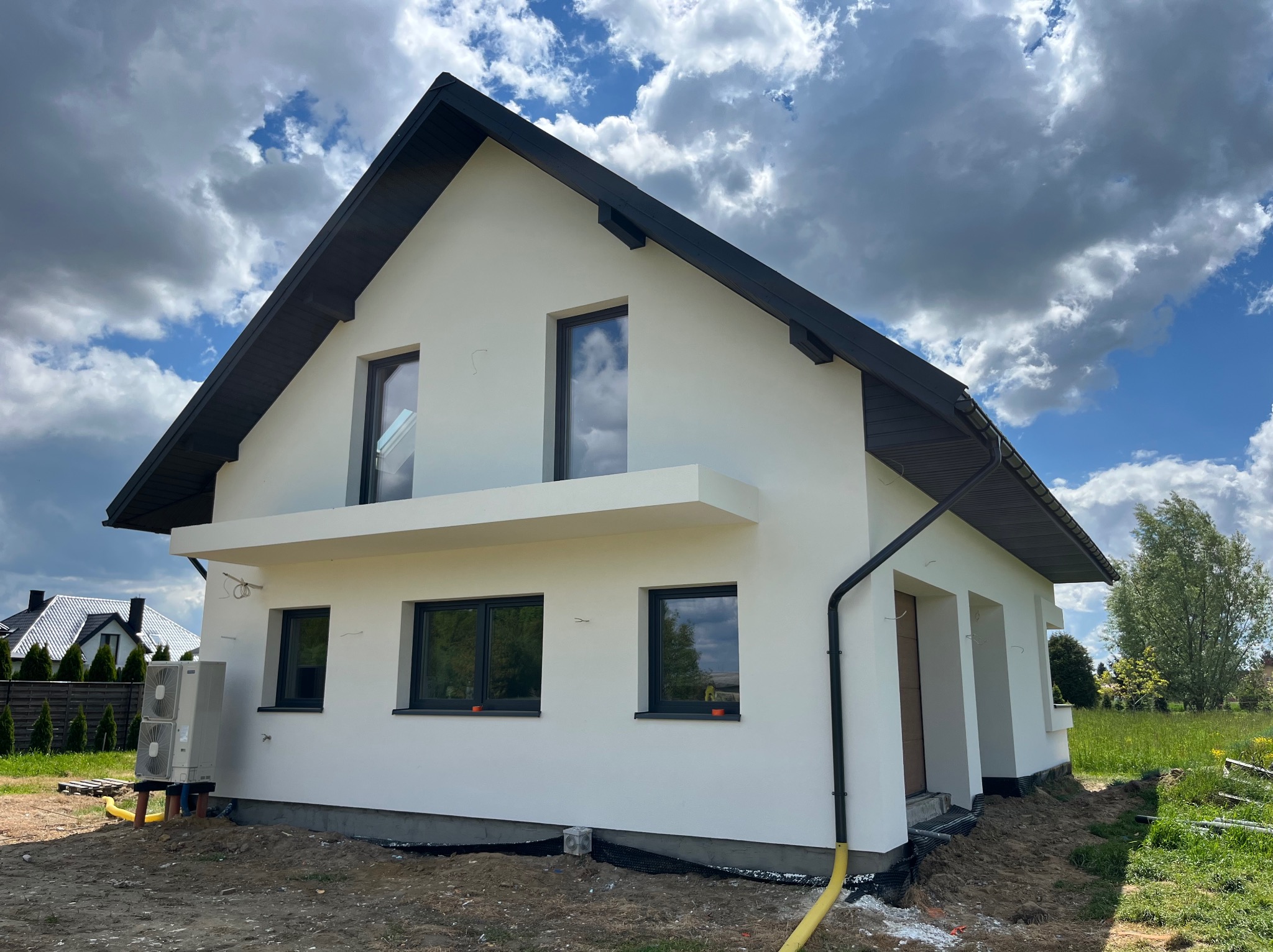 Nowoczesny dom jednorodzinny z białą elewacją i czarnym dachem na tle błękitnego nieba z chmurami. Widoczne okna z czarnymi ramami i elementy wykończeniowe.