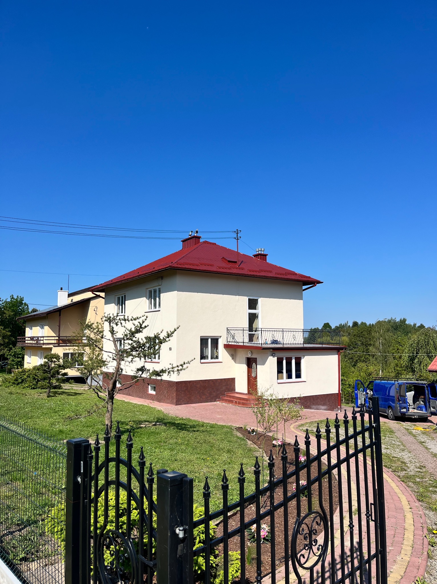 Dwupiętrowy dom z czerwonym dachem i beżową elewacją, widoczny zza czarnego, ozdobnego ogrodzenia. Na podjeździe stoi niebieski bus z otwartymi drzwiami.