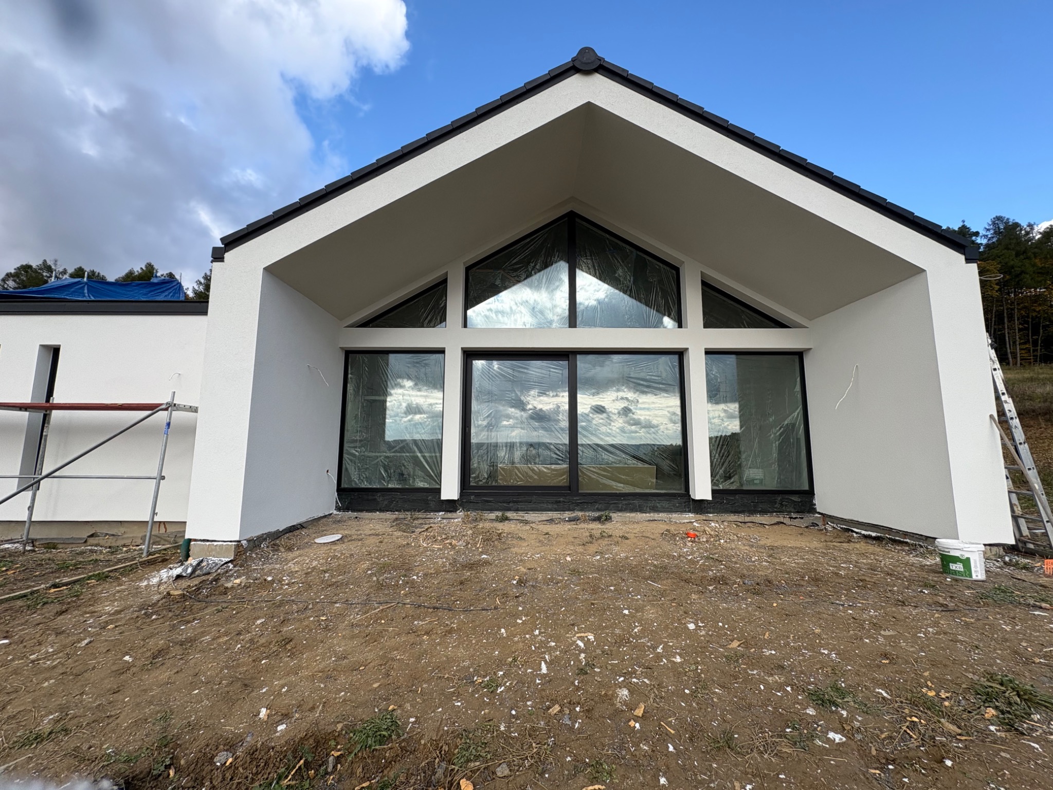Nowoczesny dom jednorodzinny z białym wykończeniem elewacji i dużymi oknami, zabezpieczonymi folią. Widoczne rusztowanie i drabina, sugerujące prace wykończeniowe.