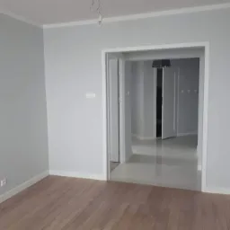 Pomalowane na szaro wnętrze mieszkania z drewnianą podłogą i widokiem na kolejne pomieszczenie z jasną, błyszczącą posadzką.