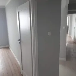 Wnętrze mieszkania po remoncie: szare ściany, białe drzwi z srebrną klamką, jasna drewniana podłoga, widok na korytarz z białymi drzwiami i jasną podłogą.