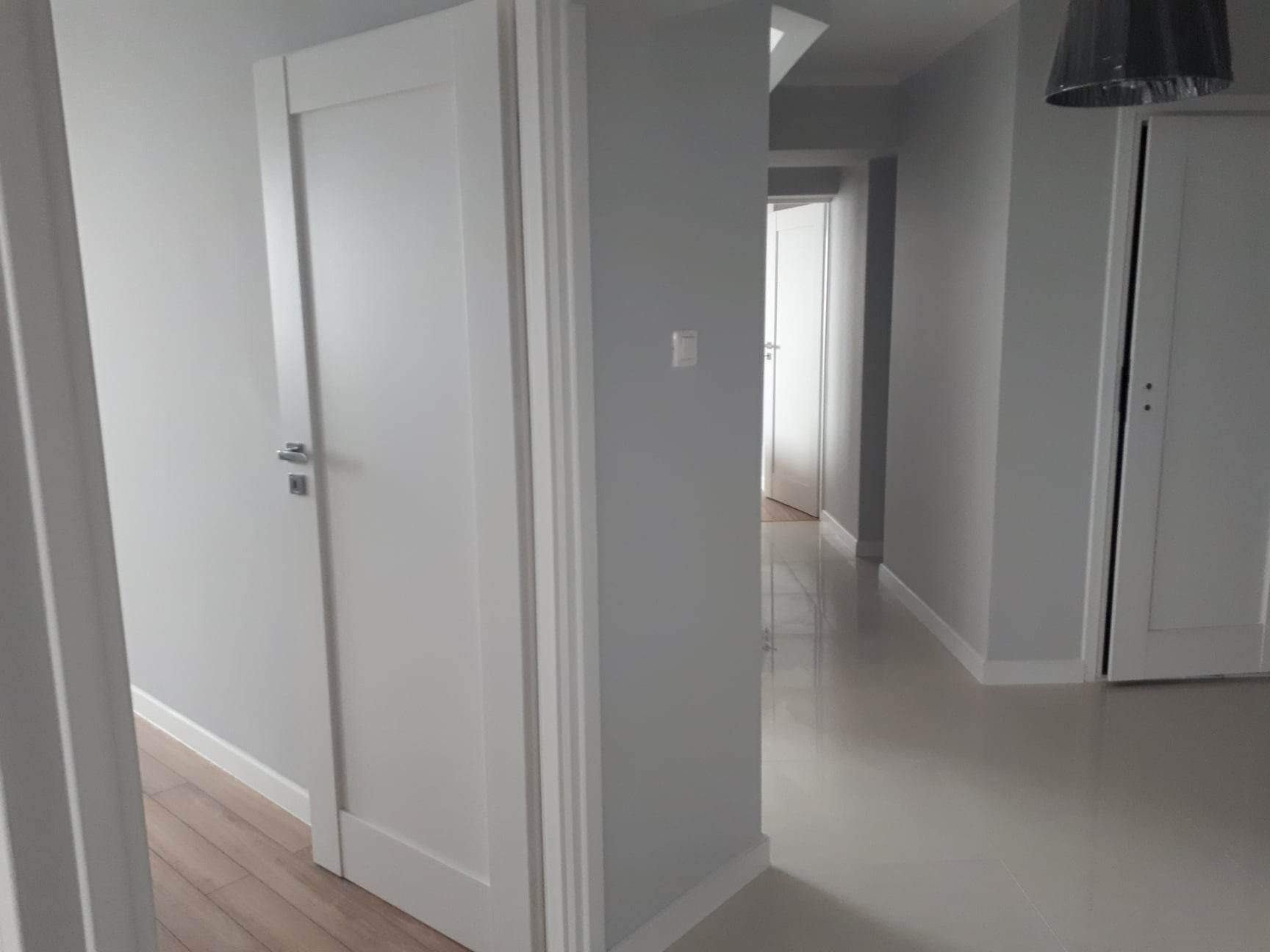 Korytarz po remoncie z szarymi ścianami, białymi drzwiami z chromowanymi klamkami i błyszczącą podłogą w jasnym odcieniu.