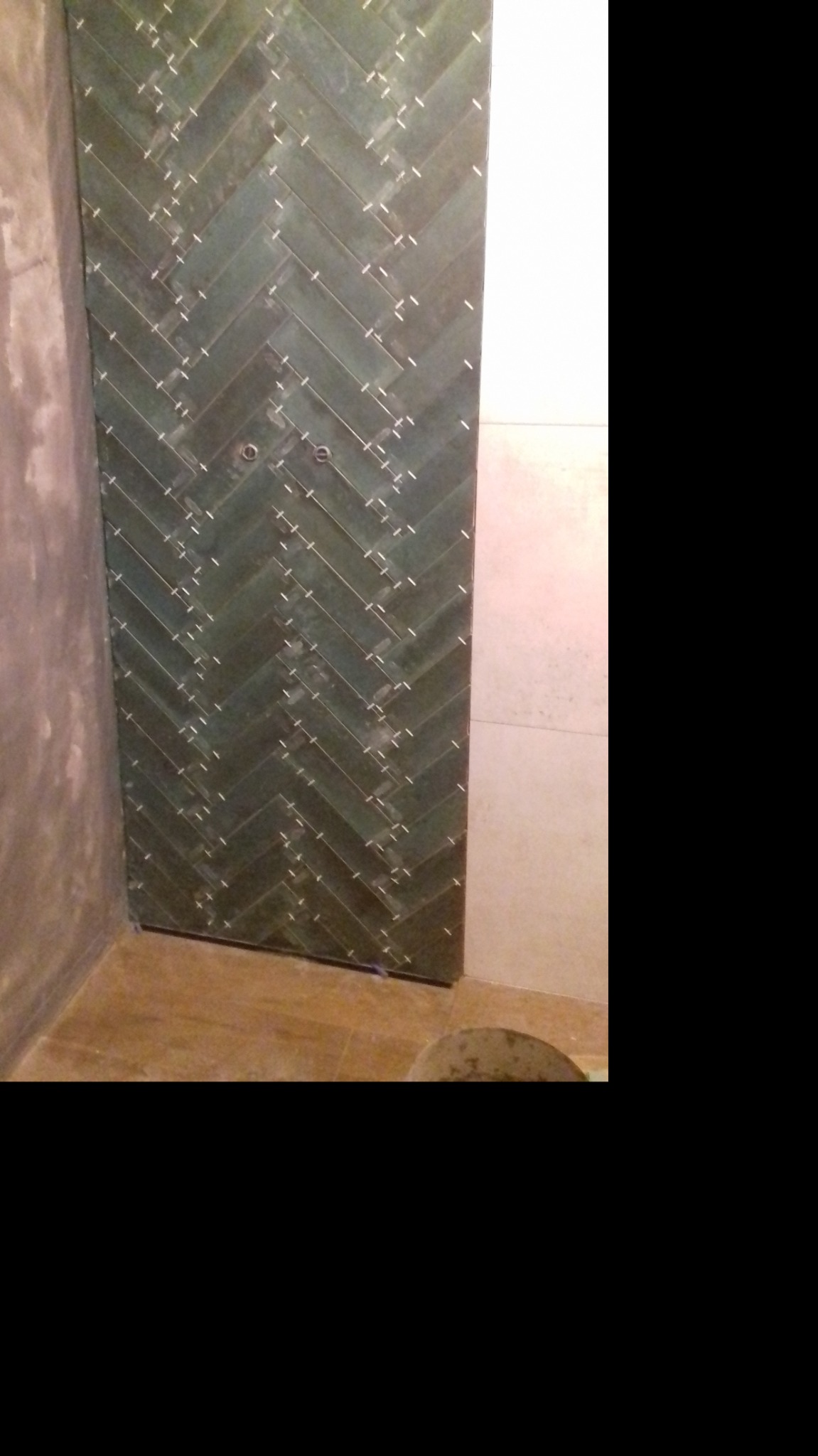 Ściana w łazience wyłożona ciemnozielonymi płytkami ułożonymi w jodełkę, z widocznymi krzyżykami dystansowymi między płytkami, w trakcie prac wykończeniowych.