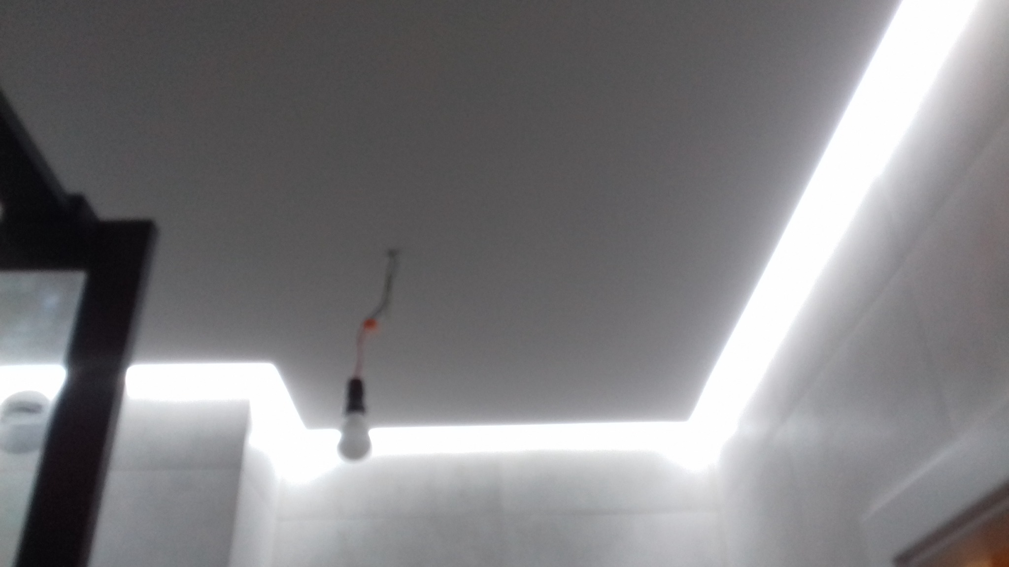Niedokończona instalacja oświetleniowa sufitu podwieszanego z widoczną żarówką na kablu i fragmentem ściany z szarymi płytkami.