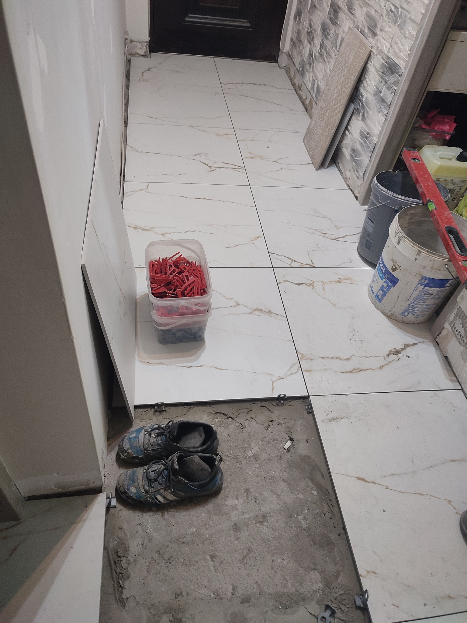 Podczas układania płytek podłogowych: widoczne białe płytki imitujące marmur, pojemnik z czerwonymi klinami dystansowymi, poziomica oparta o ścianę, oraz robocze buty przy ścianie.