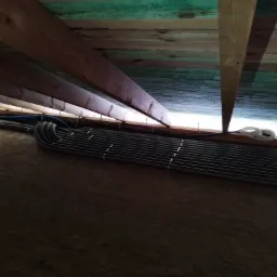 Równolegle ułożone, czarne, karbowane rury wentylacyjne przymocowane do ściany pod drewnianym stropem z widocznymi belkami i niebieskim przewodem.