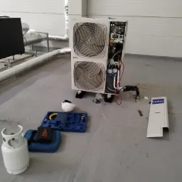 Rozłożona pompa ciepła Haier podczas serwisu w pomieszczeniu technicznym, widoczne narzędzia, butla z gazem i osłona obudowy.