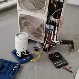 Otwarta obudowa klimatyzatora z widocznymi elementami wewnętrznymi, podłączona do manometru i butli z czynnikiem chłodniczym, obok leżą narzędzia w niebieskiej walizce i czarnym pudełku z bitami.