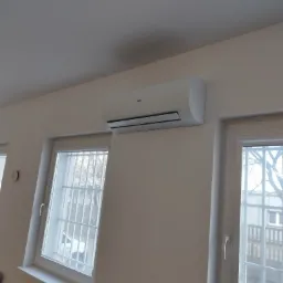 Biała jednostka klimatyzacyjna Haier zamontowana na ścianie w jasnym pomieszczeniu, widoczna pomiędzy dwoma oknami z kratami.