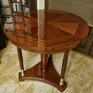 Okrągły, politurowany stolik kawowy z intarsjami w odcieniach brązu, na trójnożnej podstawie z mosiężnymi detalami, wykonany w Poznaniu. Widoczny regał z książkami w tle.