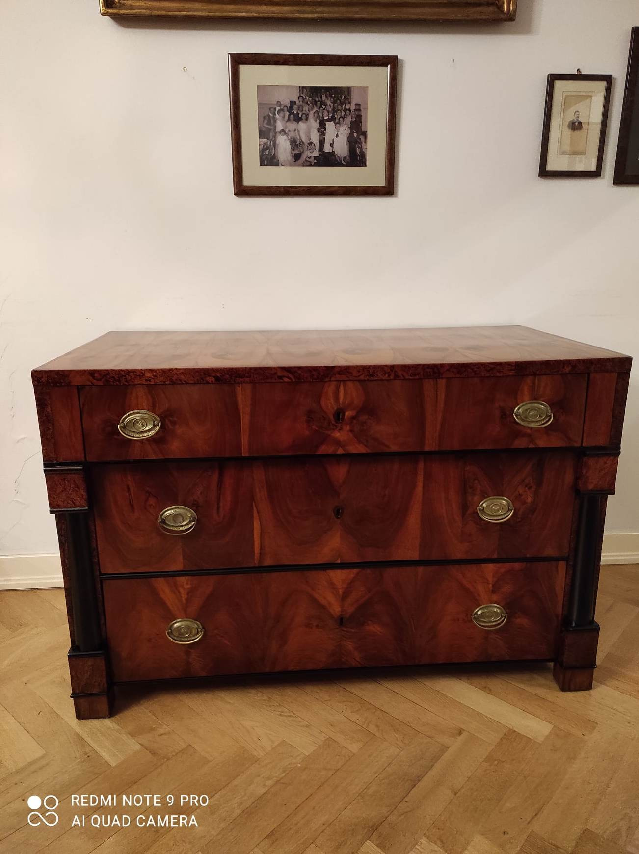 Antyczna komoda z trzema szufladami, wykonana z ciemnego drewna o wyraźnym usłojeniu, z mosiężnymi uchwytami, stojąca na parkiecie w jodełkę, na ścianie wiszą oprawione zdjęcia.