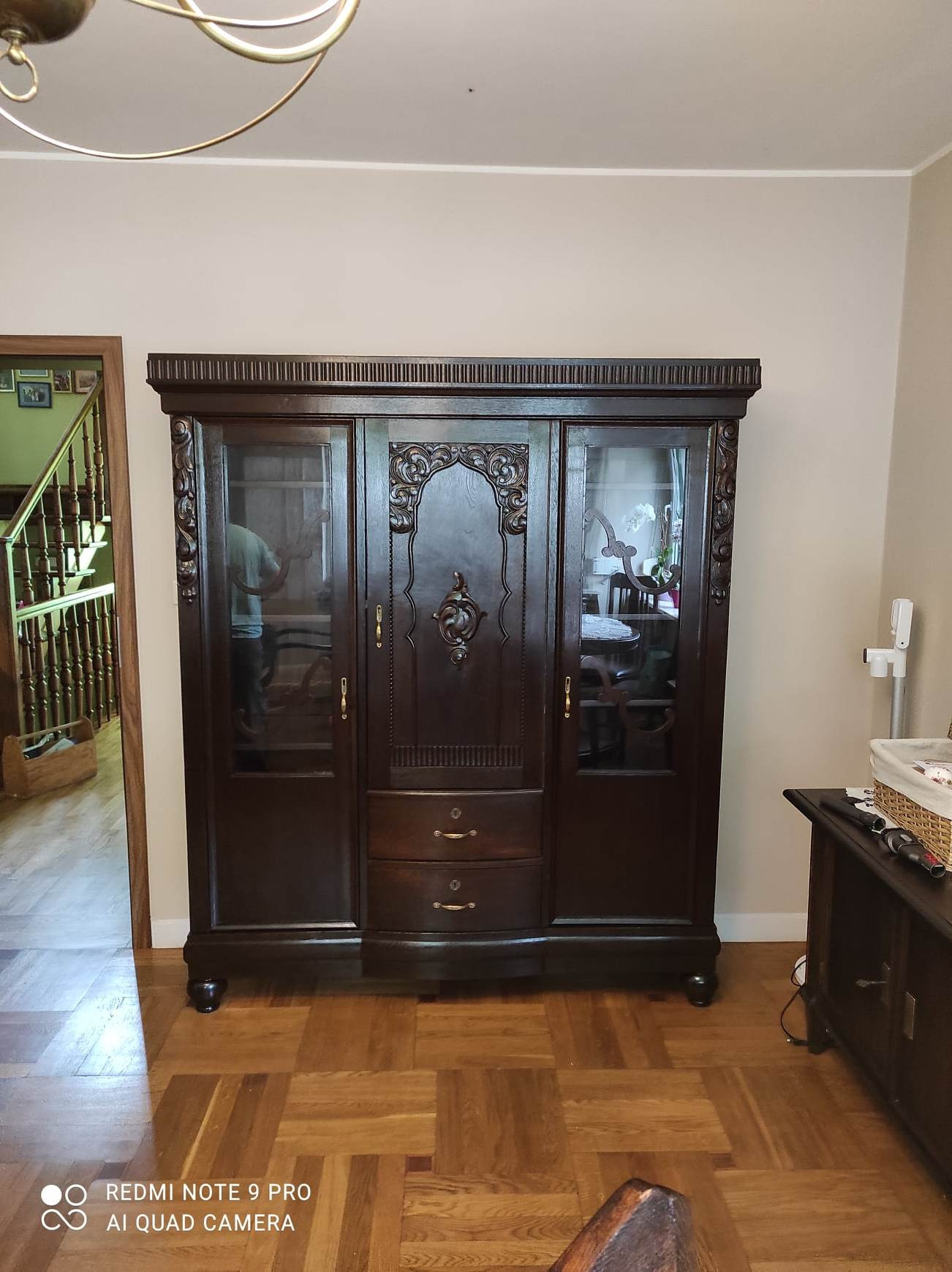 Starodawna, ciemnobrązowa szafa trzydrzwiowa z przeszklonymi witrynami i rzeźbionymi elementami dekoracyjnymi, stojąca na parkiecie w pokoju.