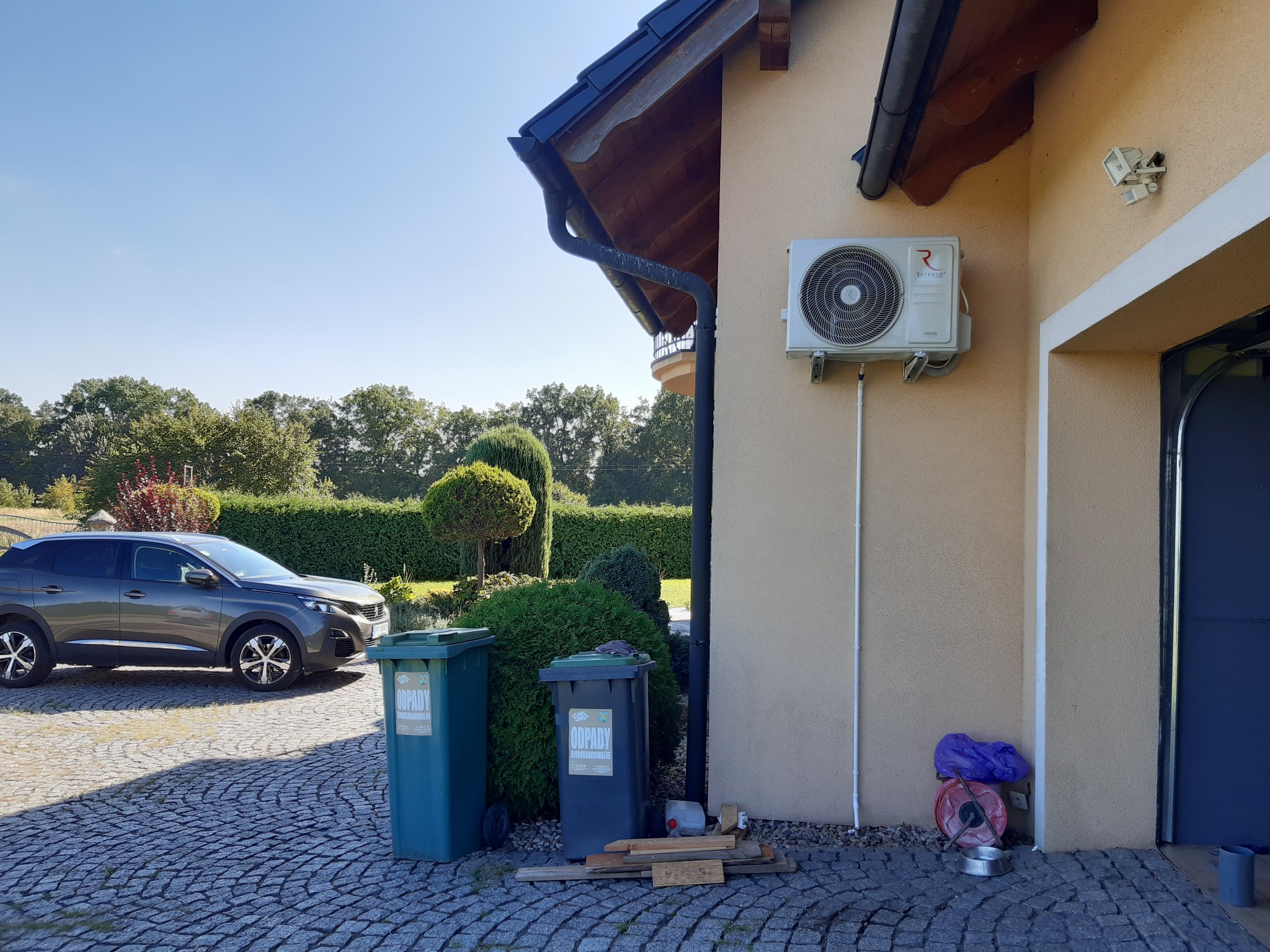 Zewnętrzna jednostka klimatyzacji Rotenso zamontowana na elewacji domu z widocznymi przewodami, obok garażu, z dwoma pojemnikami na odpady w tle.