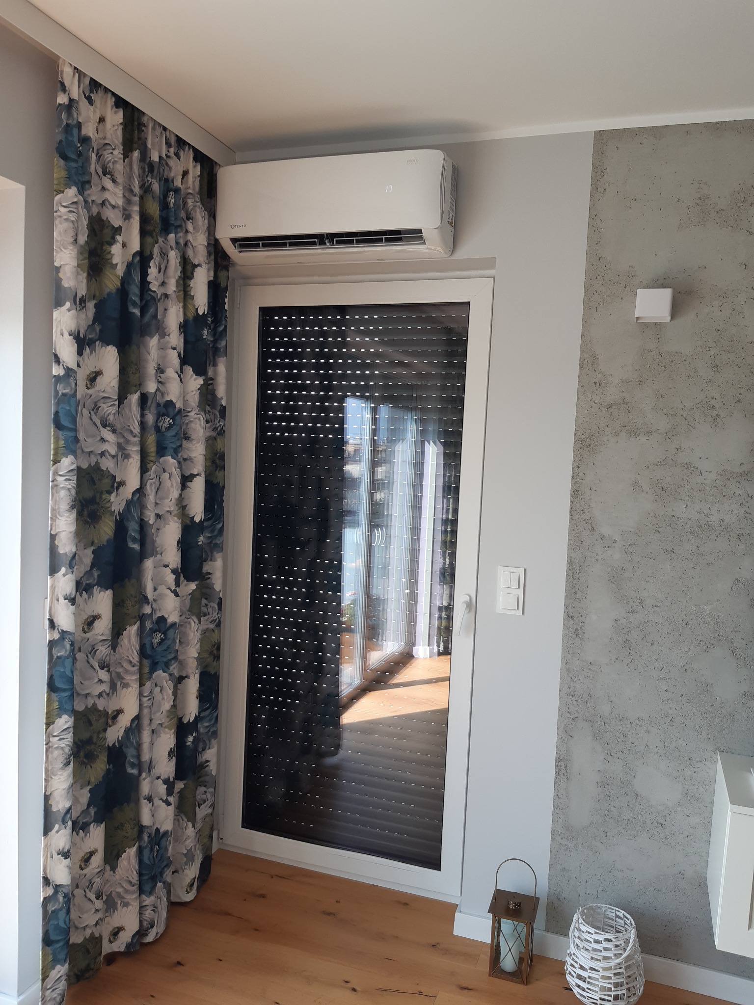 Biały klimatyzator ścienny zamontowany nad oknem z roletami wewnętrznymi w nowoczesnym salonie z drewnianą podłogą i dekoracyjnymi zasłonami w kwiaty.
