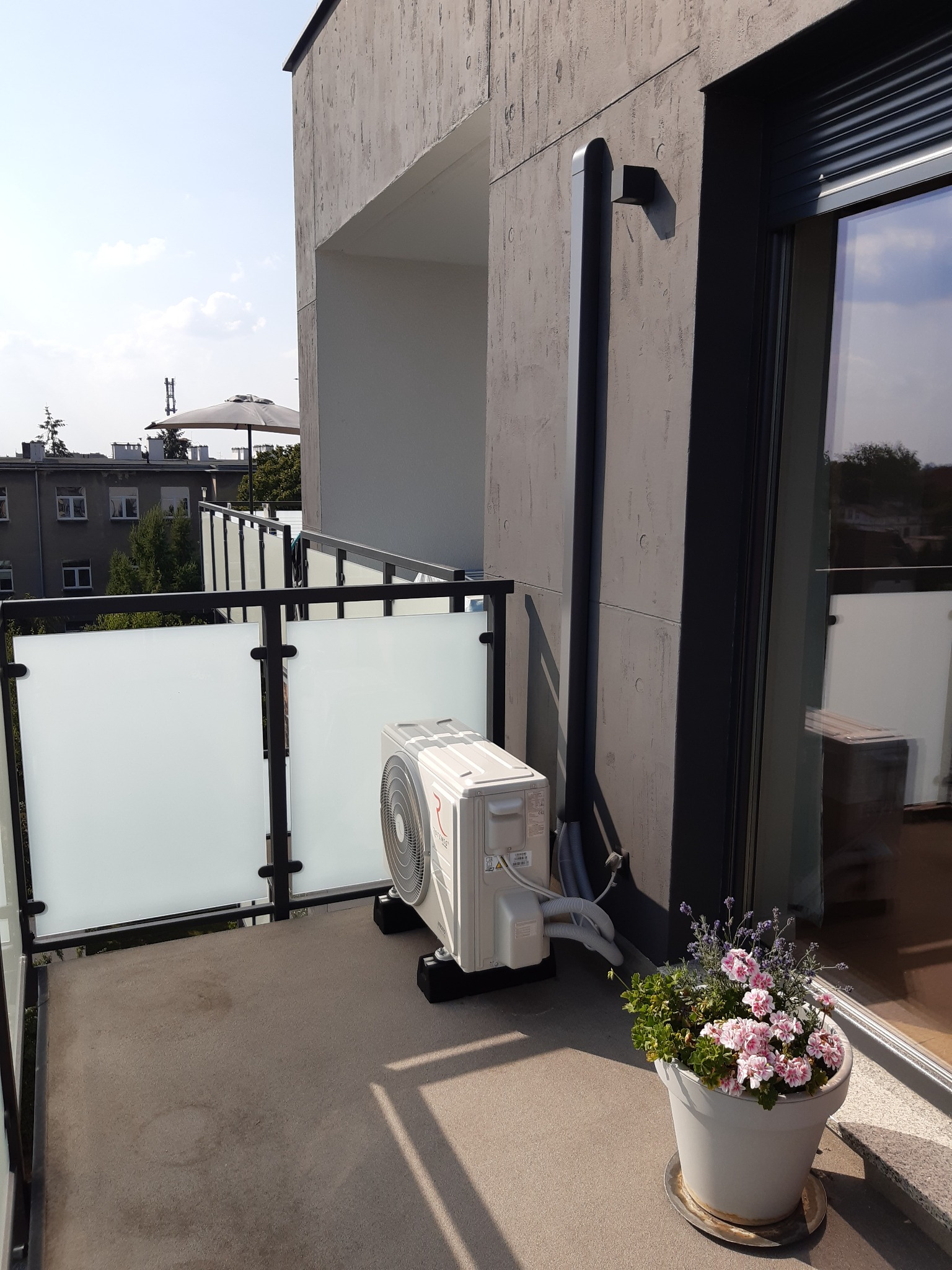 Biała jednostka zewnętrzna klimatyzacji na balkonie nowoczesnego apartamentu z widokiem na miasto, obok donica z różowymi kwiatami.