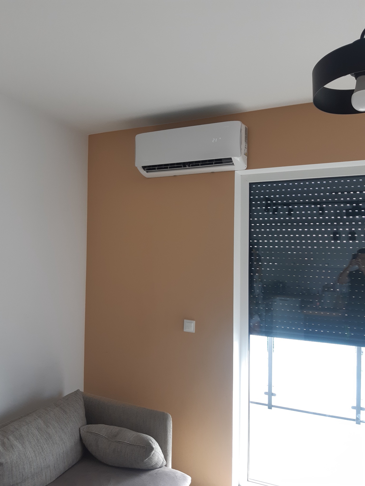 Biały klimatyzator ścienny z wyświetlaczem temperatury 21 stopni Celsjusza, zamontowany na brązowej ścianie w pokoju, widoczny fragment szarej sofy i rolety okiennej.