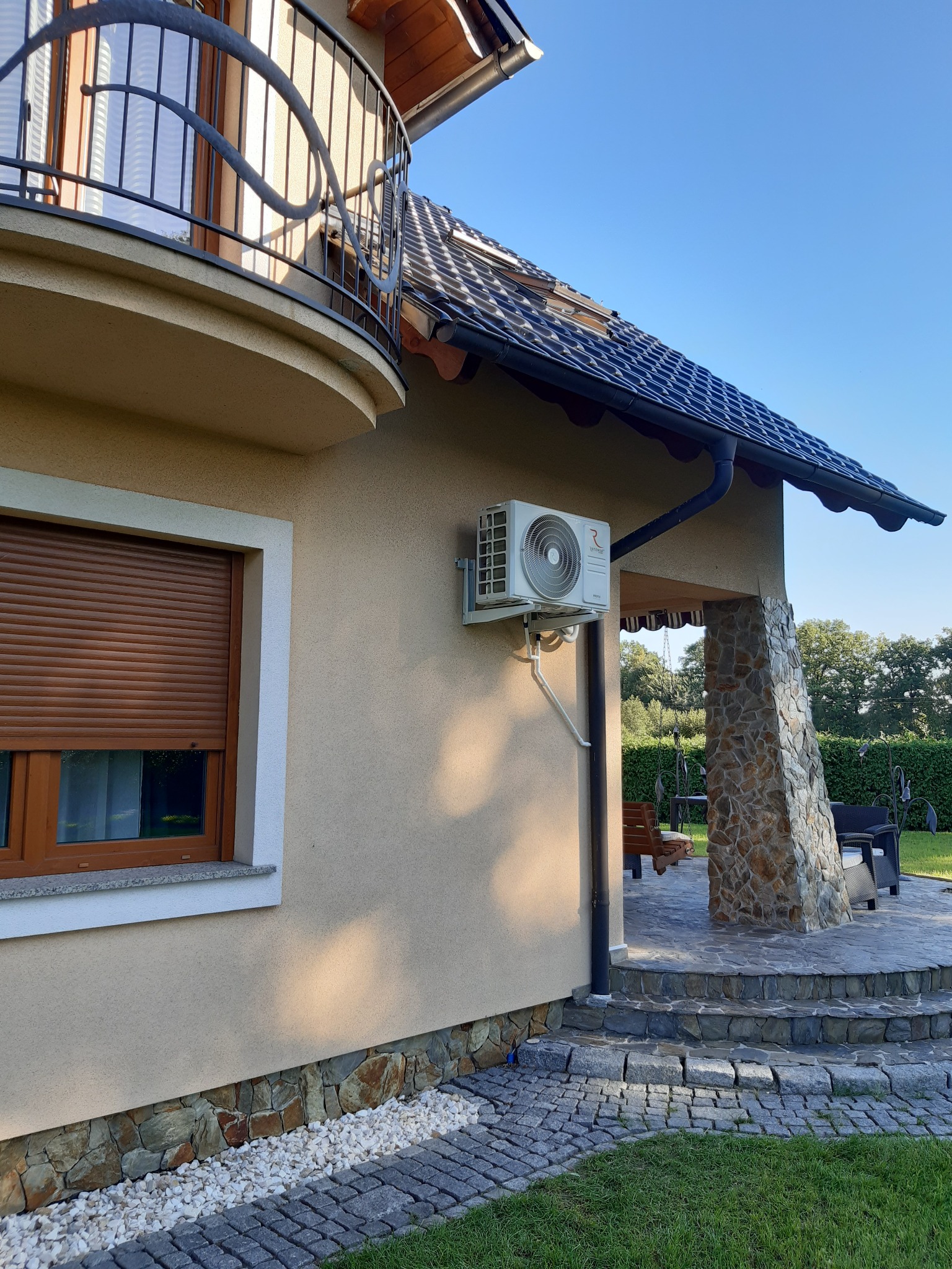 Biała jednostka zewnętrzna klimatyzatora zamontowana na elewacji beżowego domu z kamienną podmurówką i balkonem z ozdobną balustradą, widok z boku na tle niebieskiego nieba.
