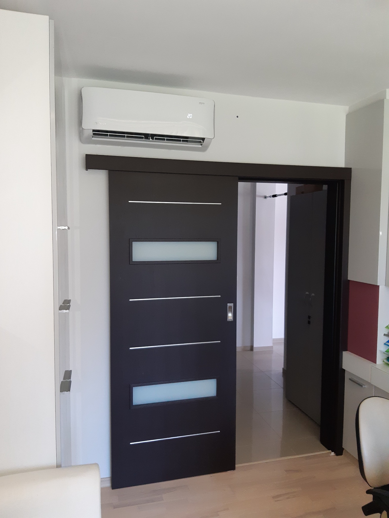 Biały klimatyzator ścienny zamontowany nad przesuwnymi, ciemnymi drzwiami z poziomymi, jasnymi paskami, widok na jasne wnętrze mieszkania.