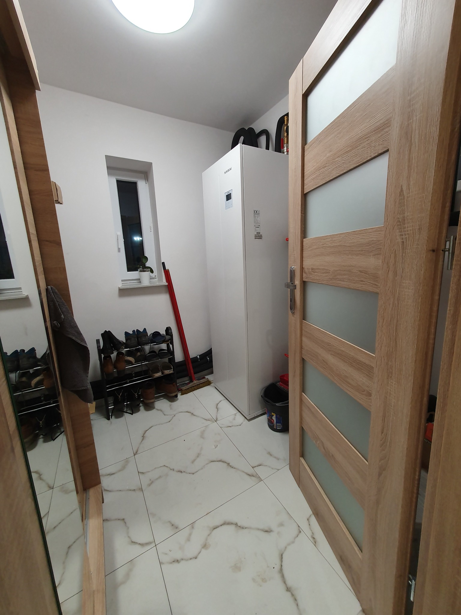 Biała jednostka wewnętrzna pompy ciepła w pomieszczeniu gospodarczym z regałem na buty, mopem i drzwiami z przeszkleniami.