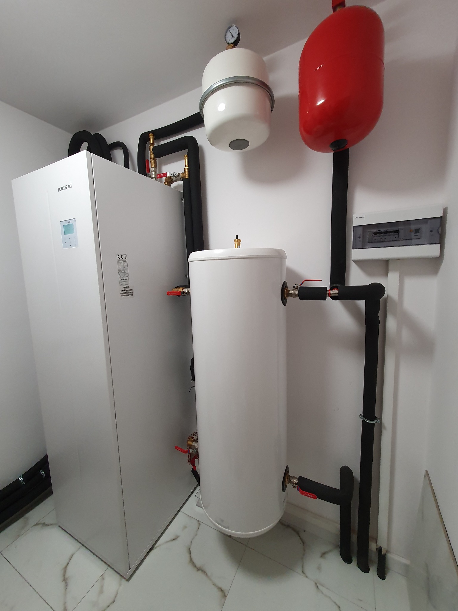 Biała pompa ciepła marki Kaisai z podłączonym zbiornikiem wody i instalacją hydrauliczną w pomieszczeniu technicznym.