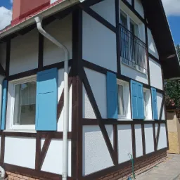 Nowoczesny dom w stylu szachulcowym z białymi ścianami, brązowymi belkami i błękitnymi okiennicami, widok z boku na elewację i fragment dachu z kominem, obok domu ułożona kostka brukowa.