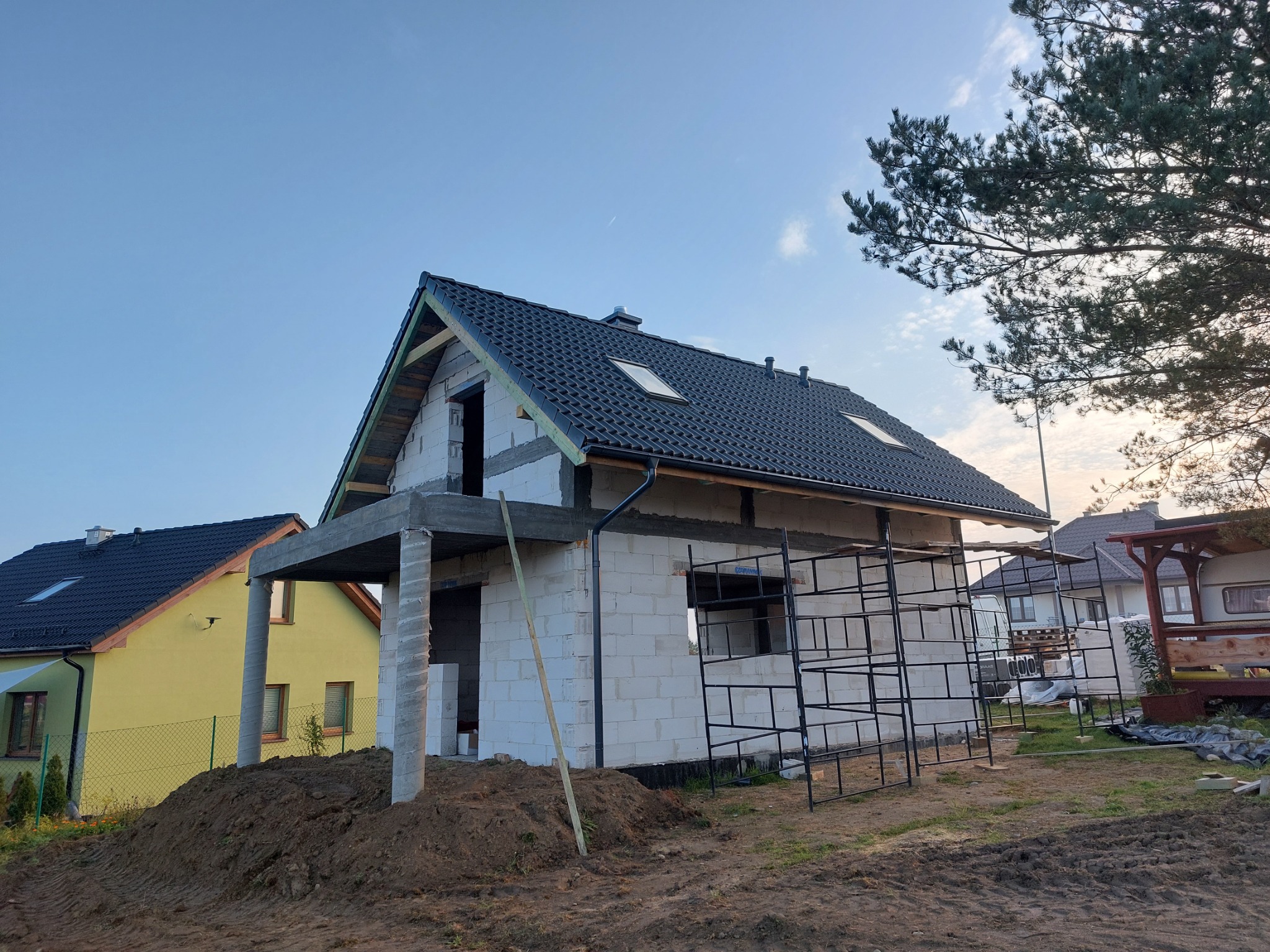 Dom murowany w stanie surowym zamkniętym z czarną dachówką, widoczne rusztowania i przygotowany teren pod ogród.