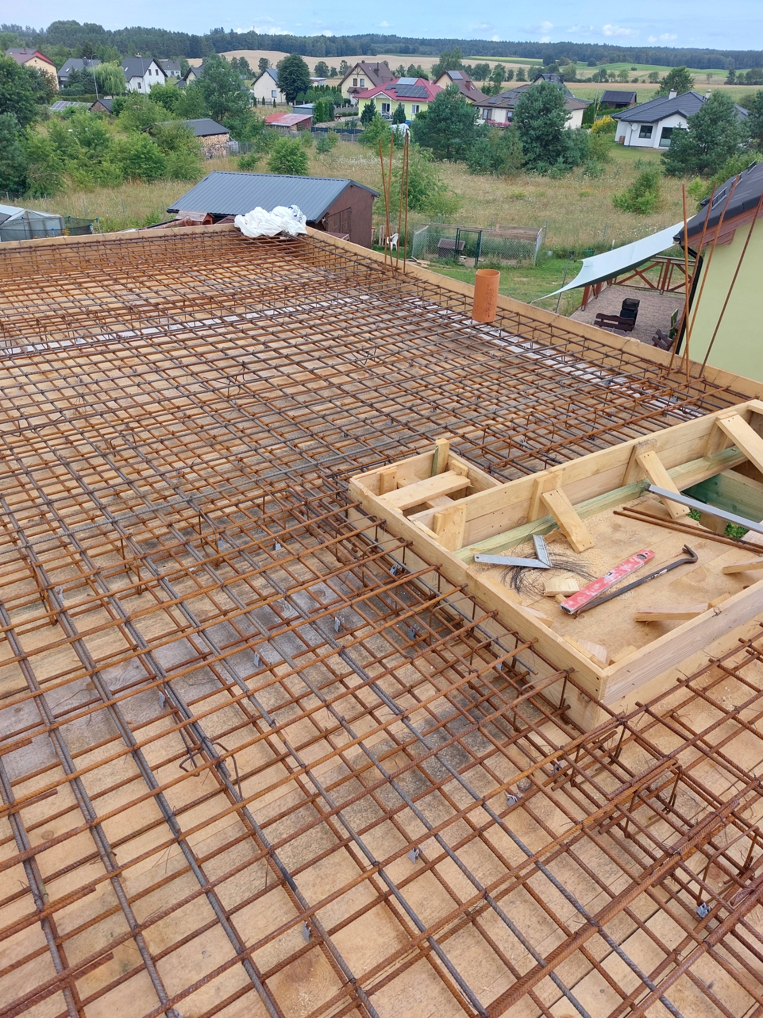 Zbrojenie stropu z drewnianym szalunkiem i narzędziami na tle zabudowy jednorodzinnej.