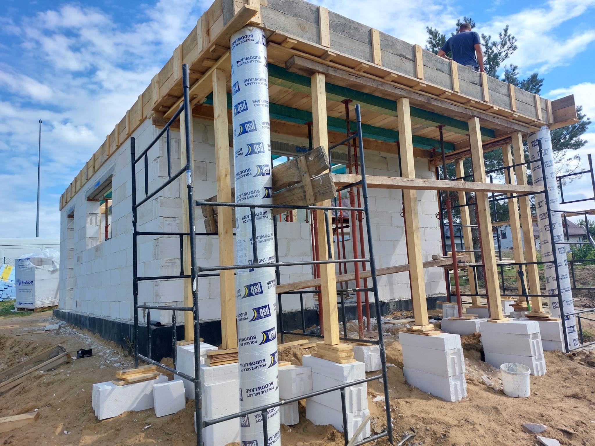 Budowa domu z białych bloczków: widoczne ściany w trakcie wznoszenia, drewniany szalunek na stropie, rusztowania i materiały budowlane na placu budowy, pracownik na górze konstrukcji.