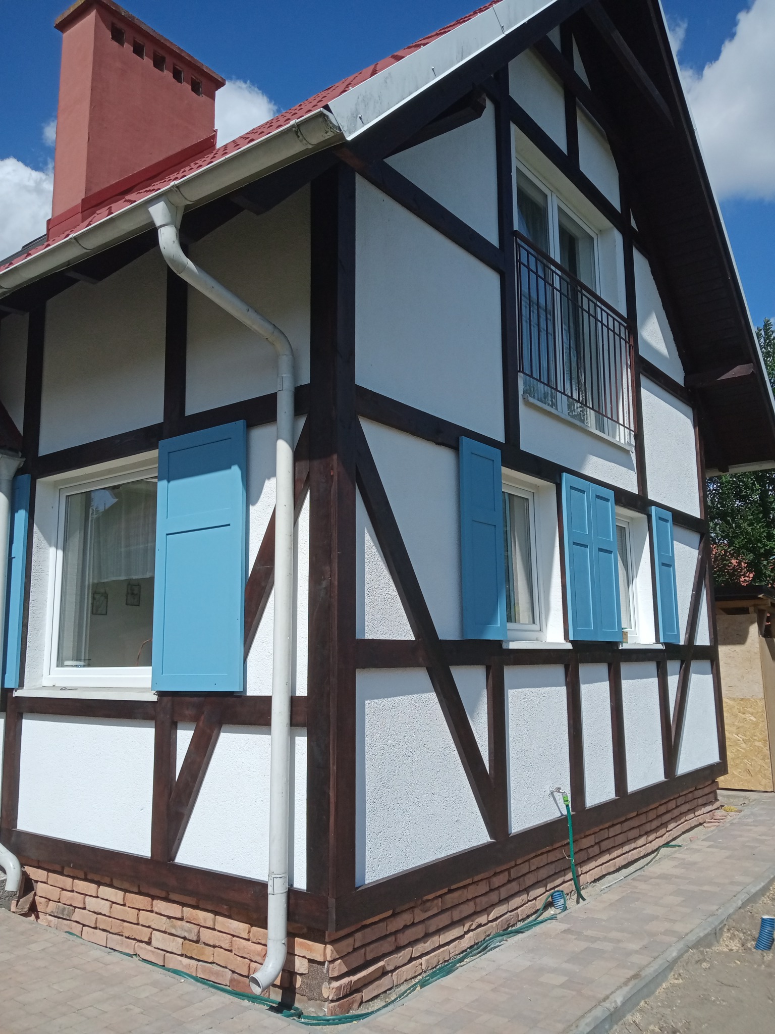 Nowoczesny dom w stylu szachulcowym z białymi ścianami, brązowymi belkami i błękitnymi okiennicami, widok z boku na elewację i fragment dachu z kominem, obok domu ułożona kostka brukowa.