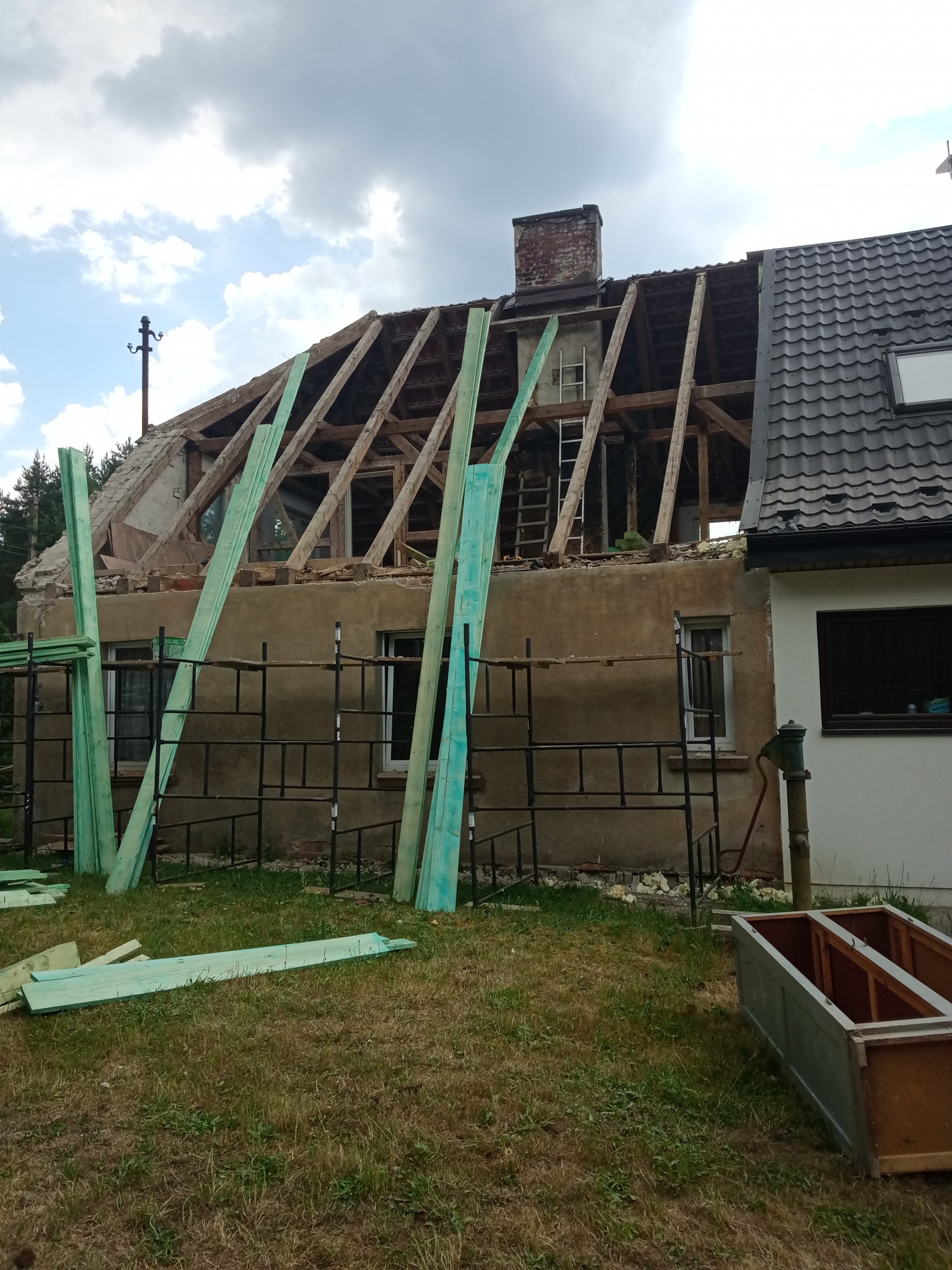 Dom w trakcie remontu dachu: odsłonięta więźba dachowa, rusztowania przy elewacji, przygotowane deski w kolorze morskim, widoczny komin z cegły i fragment nowego pokrycia dachowego.