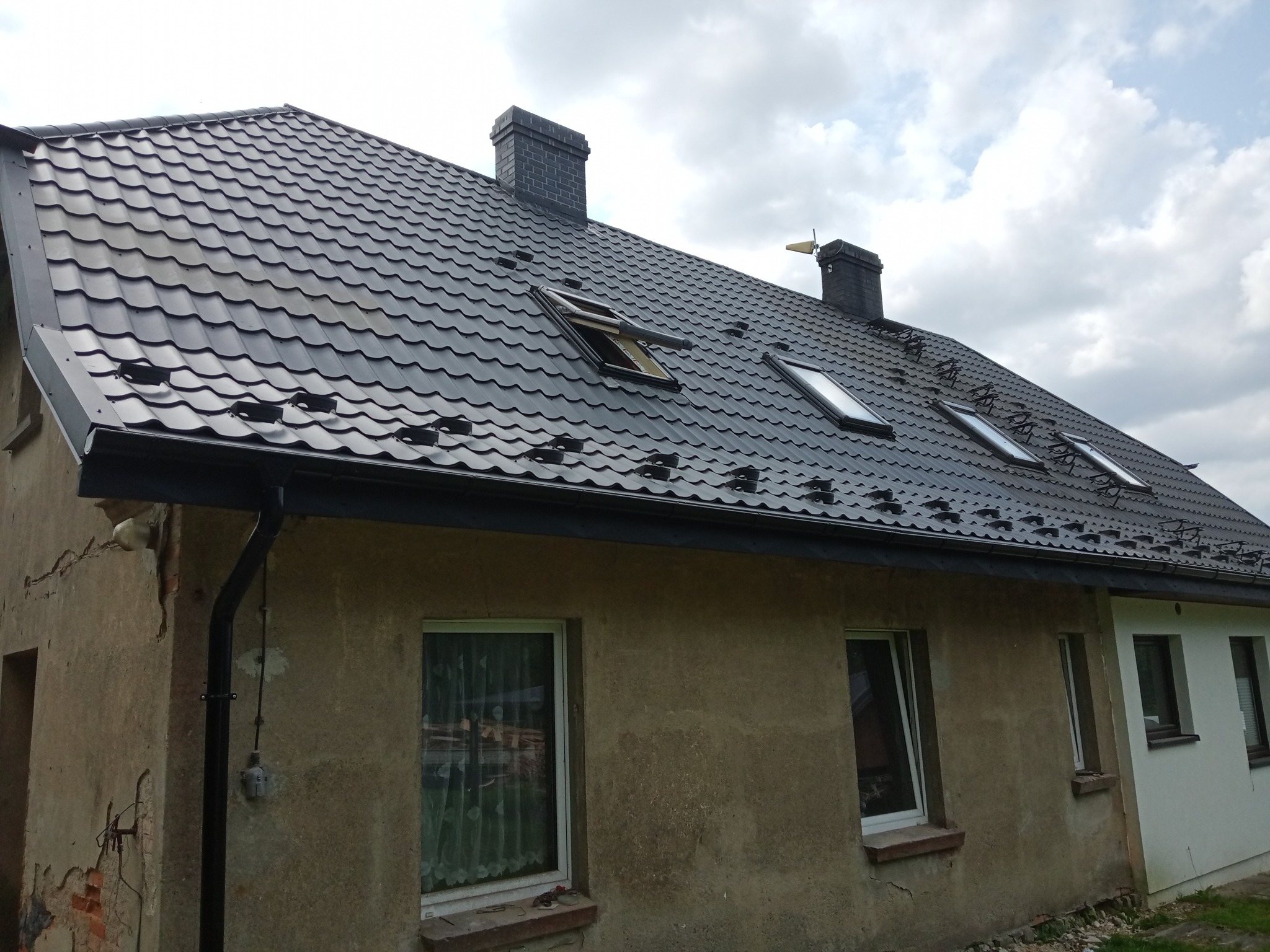 Dom z nowym, ciemnym dachem z dachówki, rynnami i oknami dachowymi, część elewacji odnowiona, a część w stanie surowym.