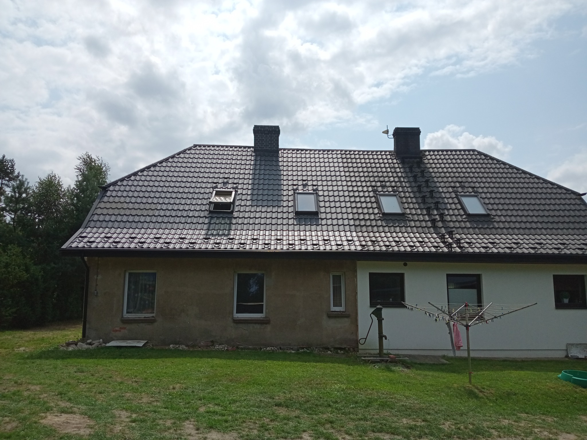 Dwukondygnacyjny dom z ciemną dachówką, widoczne kominy, okna dachowe i fragment elewacji w dwóch kolorach - szarym i białym, na trawniku rozłożona suszarka na pranie.