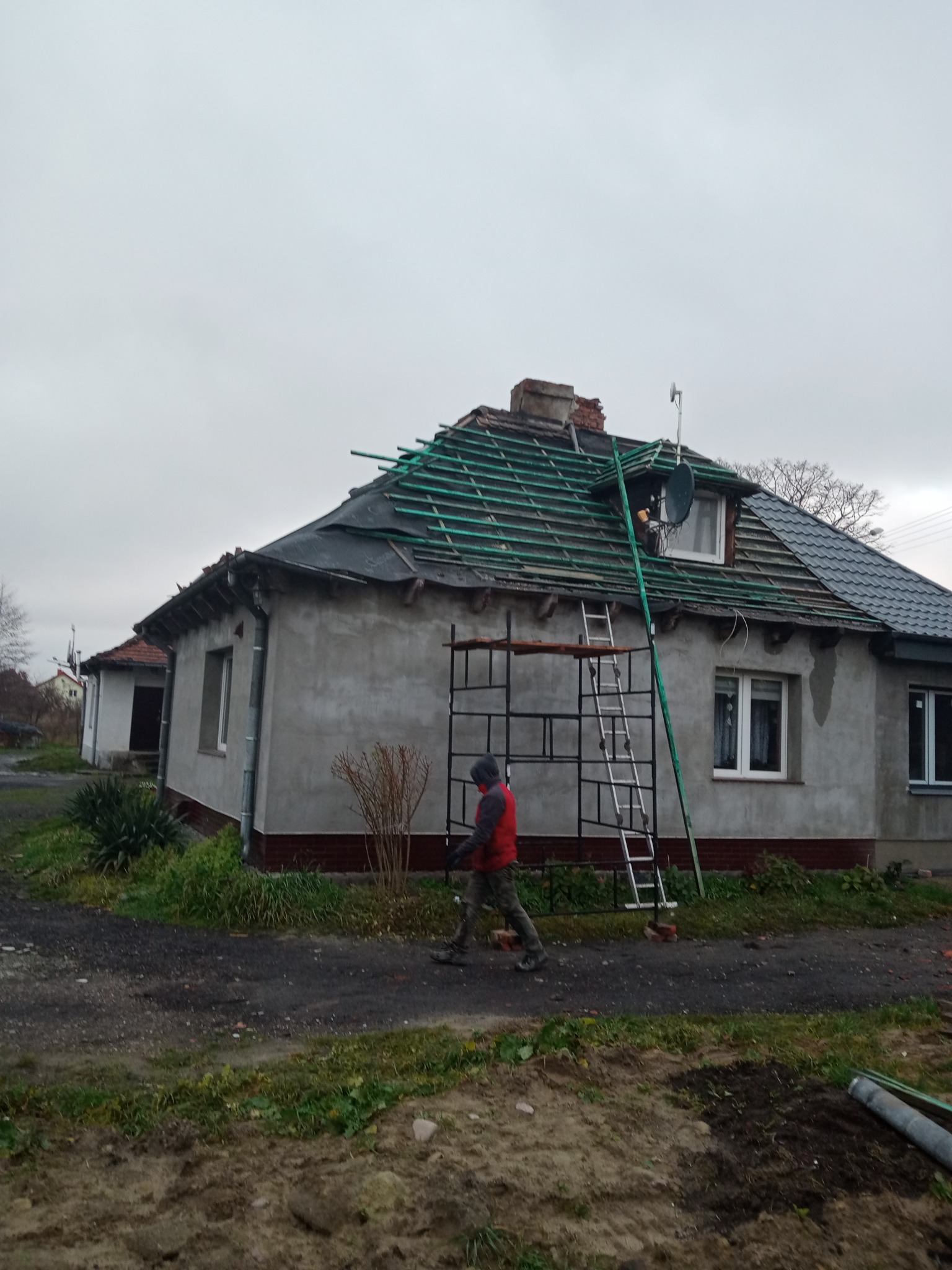 Dom jednorodzinny w trakcie wymiany pokrycia dachowego, widoczne rusztowanie i drabina oparta o dach, część dachu pokryta nową dachówką, a część przygotowana pod nowe pokrycie z widocznymi łatami.