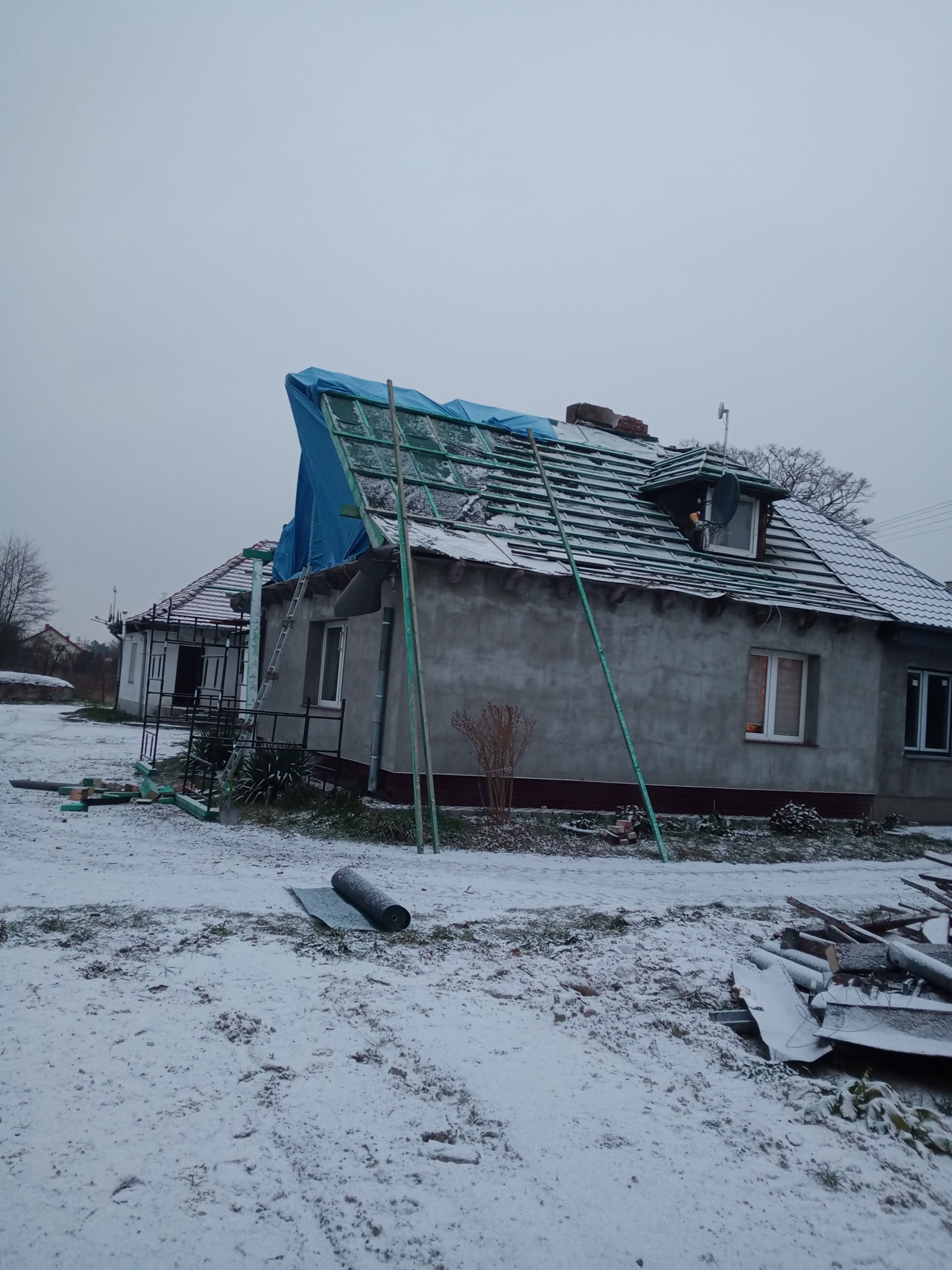 Dom jednorodzinny w trakcie remontu dachu, zabezpieczony niebieską plandeką, z widocznymi elementami konstrukcyjnymi i śniegiem na dachu oraz w otoczeniu.
