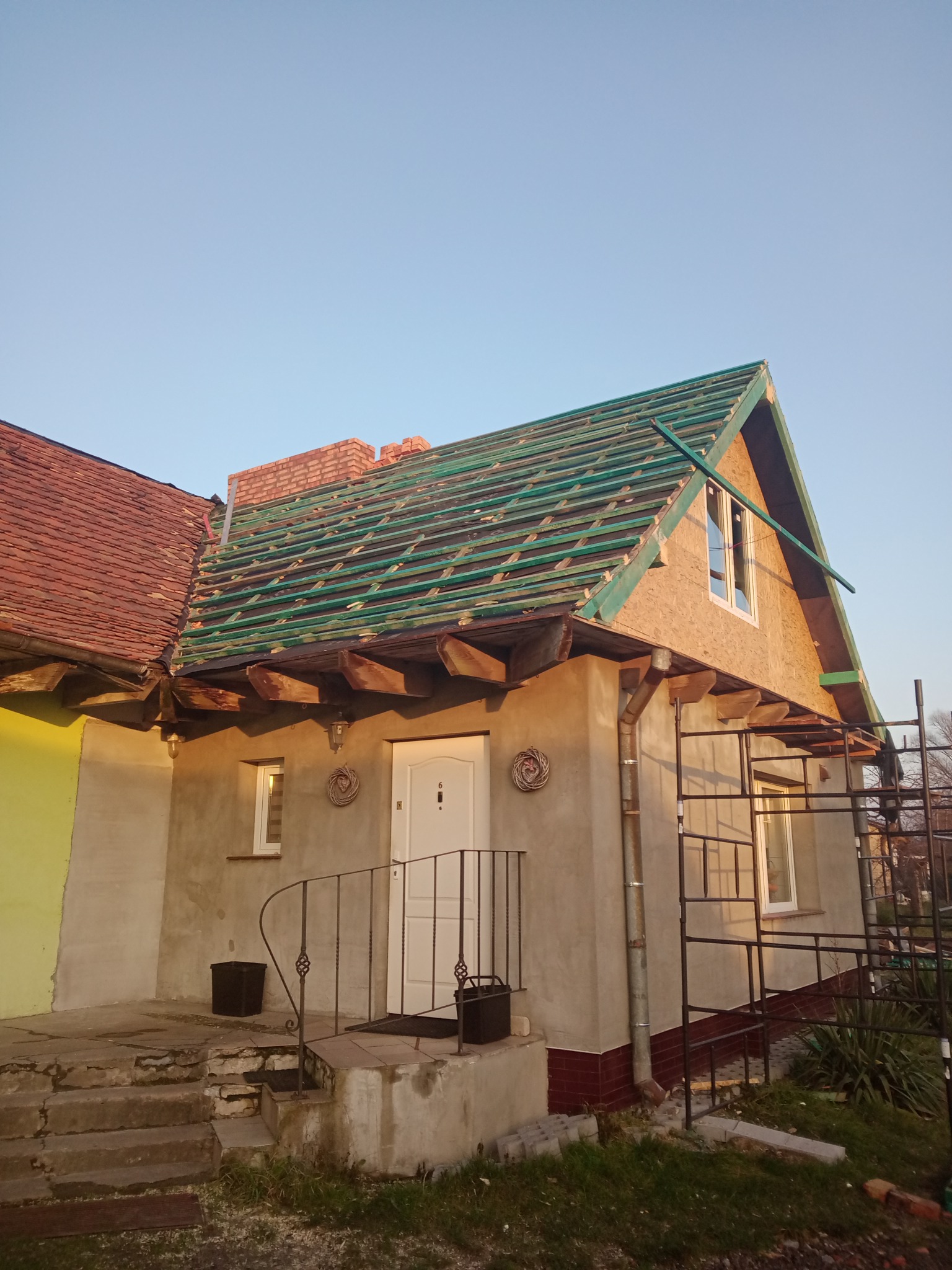 Dom w trakcie remontu dachu: stara dachówka z jednej strony, nowa więźba dachowa z łatami z drugiej strony, widoczne rusztowanie i elewacja w trakcie prac.