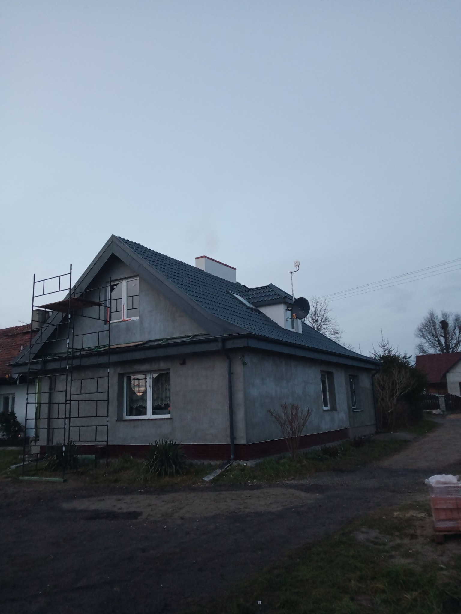 Szary dom jednorodzinny z ciemną dachówką, częściowo pokryty rusztowaniem, w trakcie prac wykończeniowych elewacji, widoczny fragment ulicy.