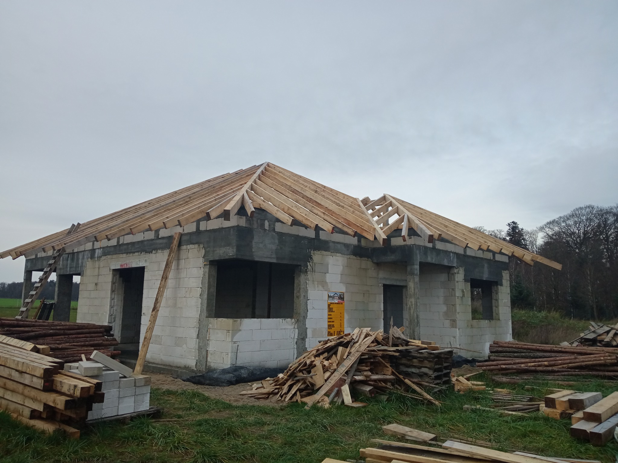 Budowa domu murowanego w stanie surowym otwartym, widoczne ściany z bloczków betonowych, drewniana konstrukcja dachu bez pokrycia, stosy drewna budowlanego na pierwszym planie, pochmurne niebo w tle.