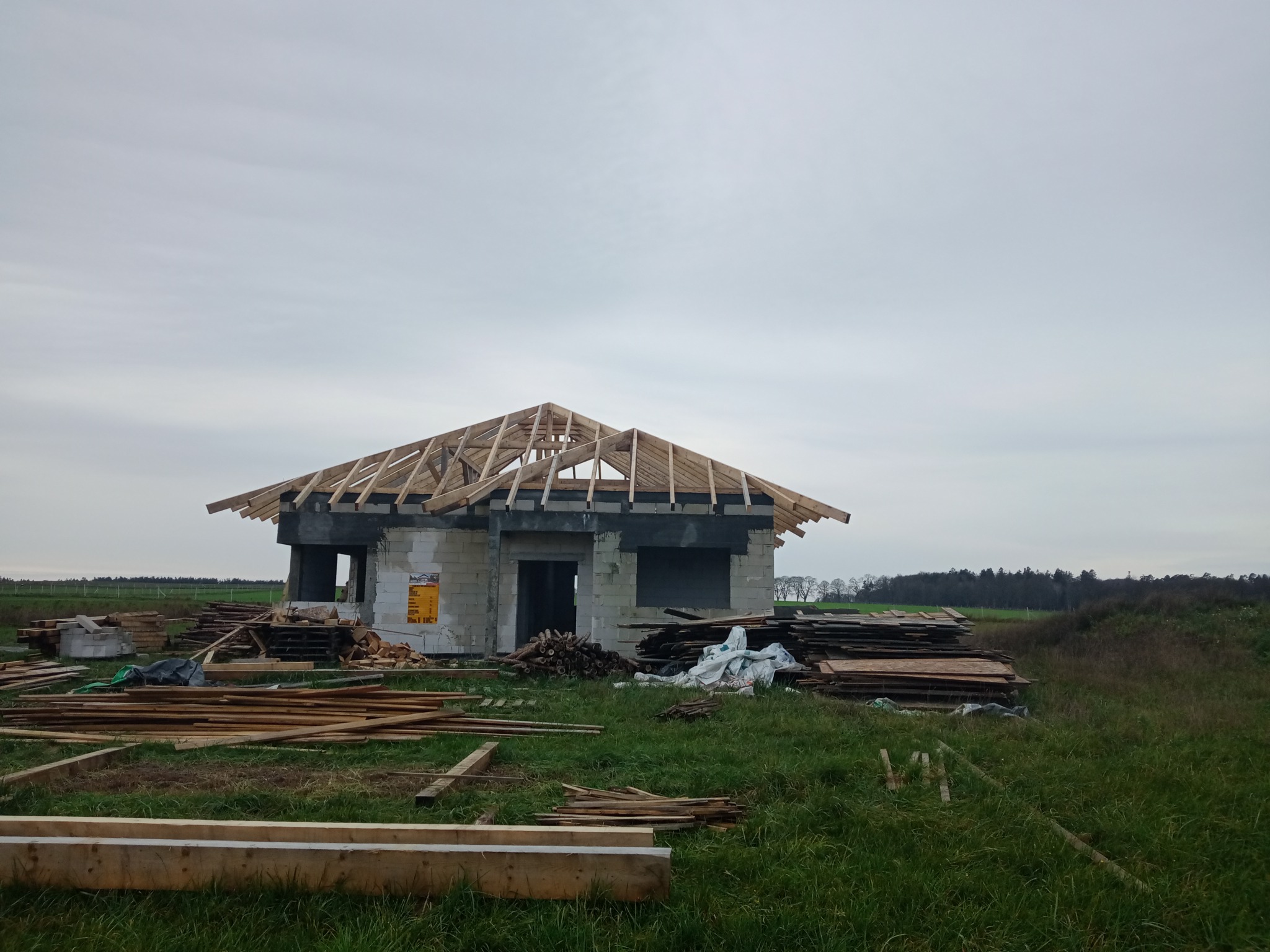 Surowy stan domu murowanego z widoczną konstrukcją dachu, otoczonego zielonym terenem i stertami drewna.