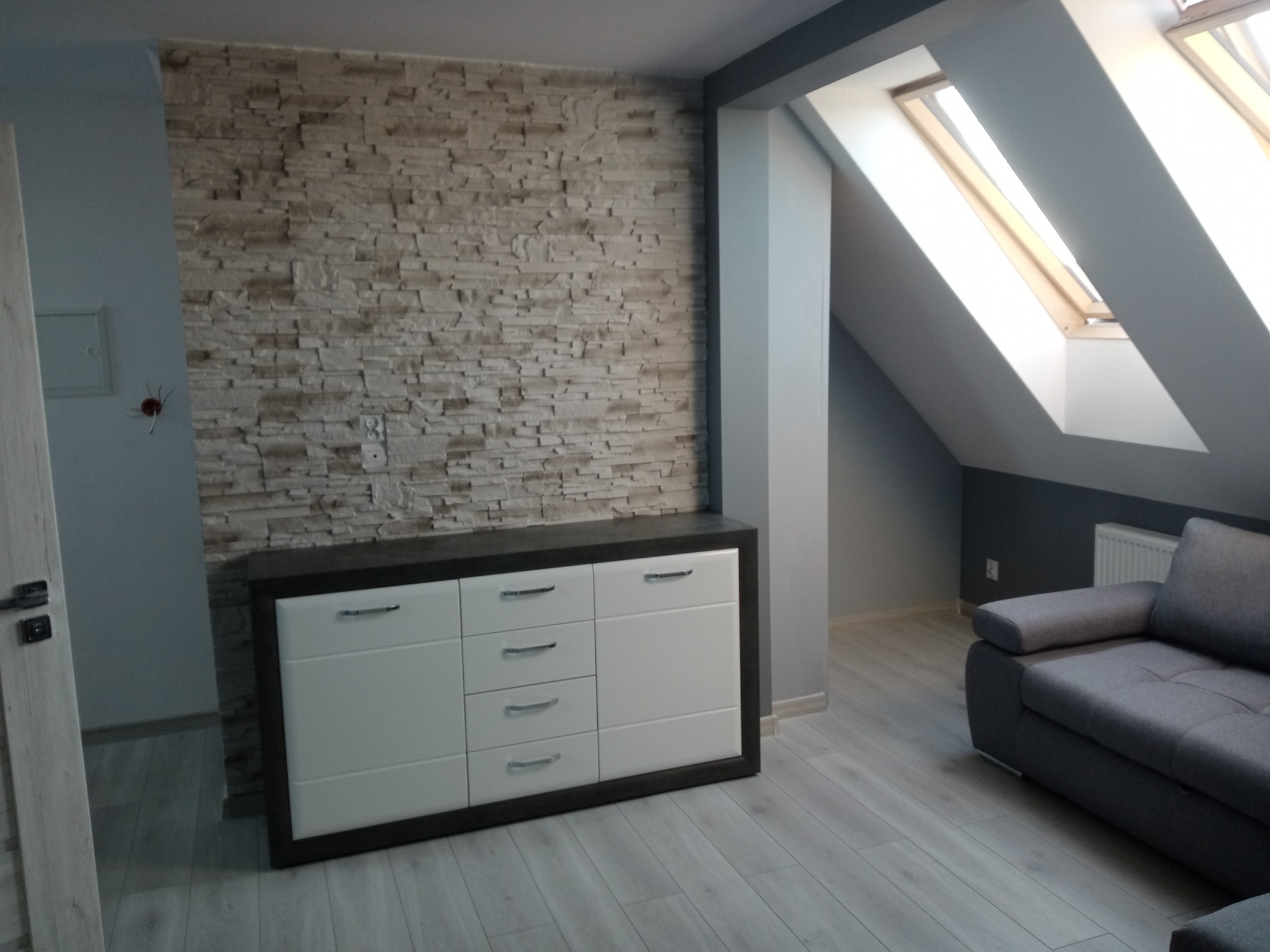 Jasne wnętrze poddasza z dekoracyjną ścianą z kamienia, białą komodą z ciemnym blatem i szarą sofą; podłoga wyłożona panelami w odcieniach szarości, a w tle widoczne okno dachowe.