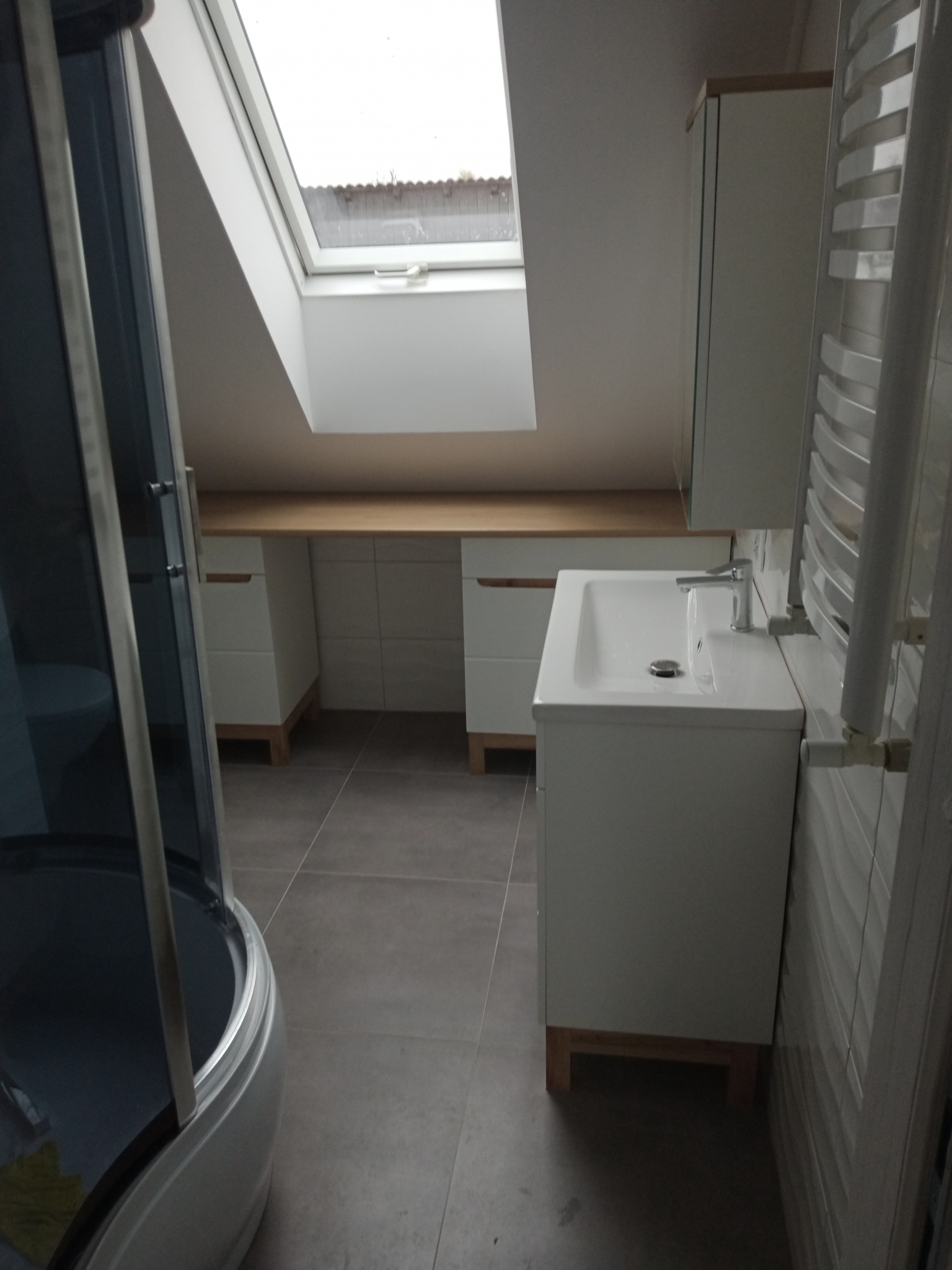 Nowoczesna łazienka na poddaszu z kabiną prysznicową, białą umywalką z szafką, grzejnikiem drabinkowym i oknem dachowym. Szare płytki podłogowe i białe ściany tworzą minimalistyczny wystrój.