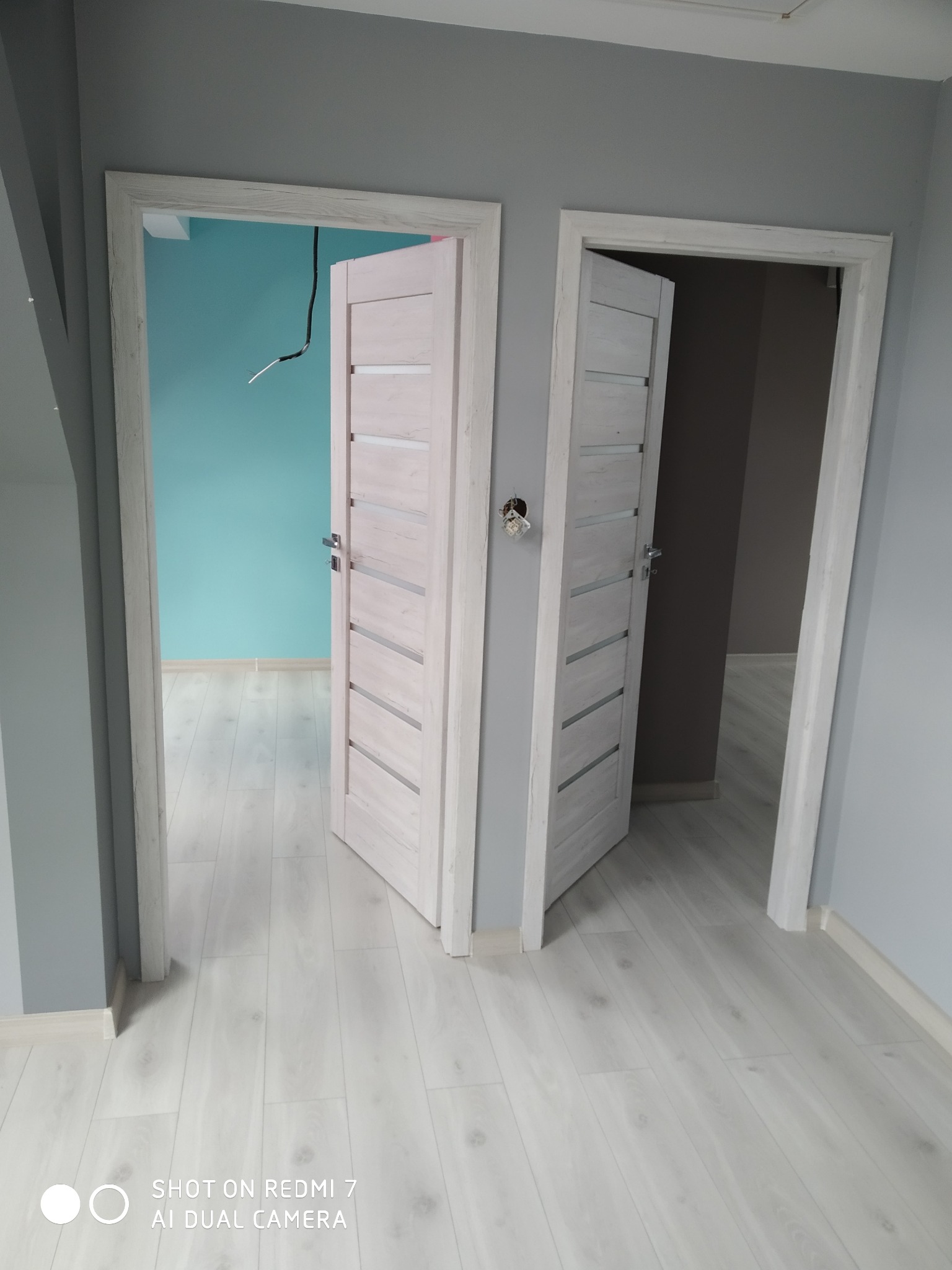 Dwa otwarte, białe drzwi wewnętrzne z przeszkleniami, widok na pomalowane ściany w odcieniach turkusu i szarości, na podłodze panele w jasnym odcieniu drewna.