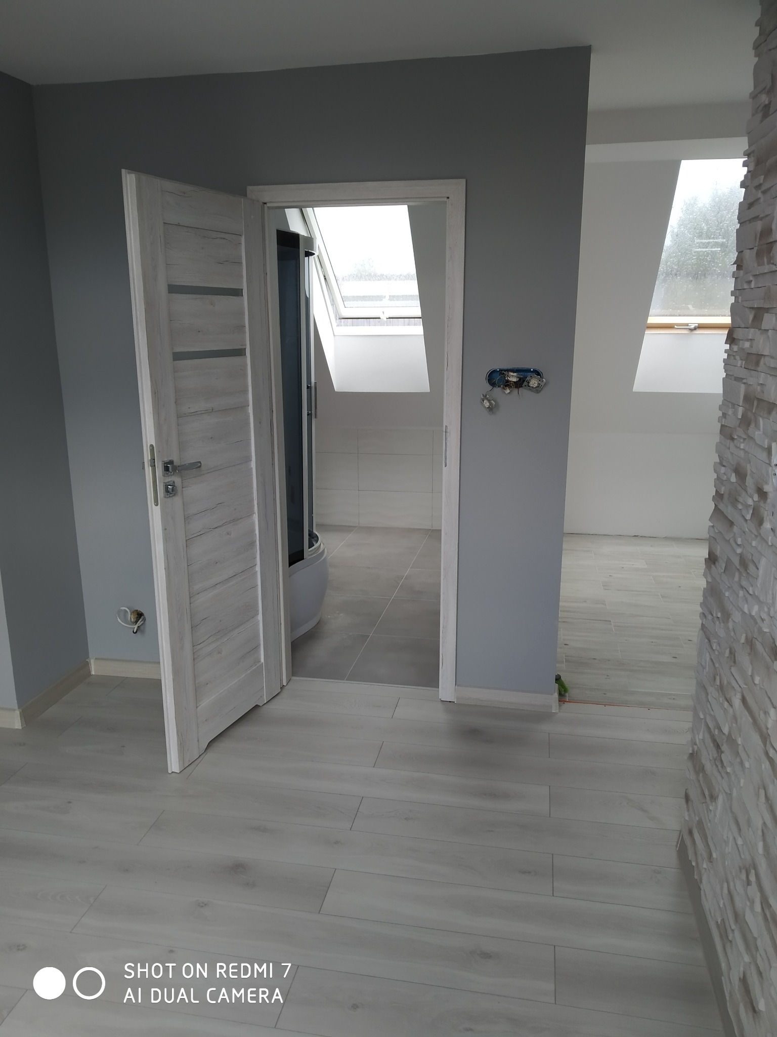 Wnętrze po remoncie: Szare ściany, jasna podłoga imitująca drewno, otwarte drzwi do łazienki z kabiną prysznicową i oknem dachowym, fragment dekoracyjnej ściany z kamienia.