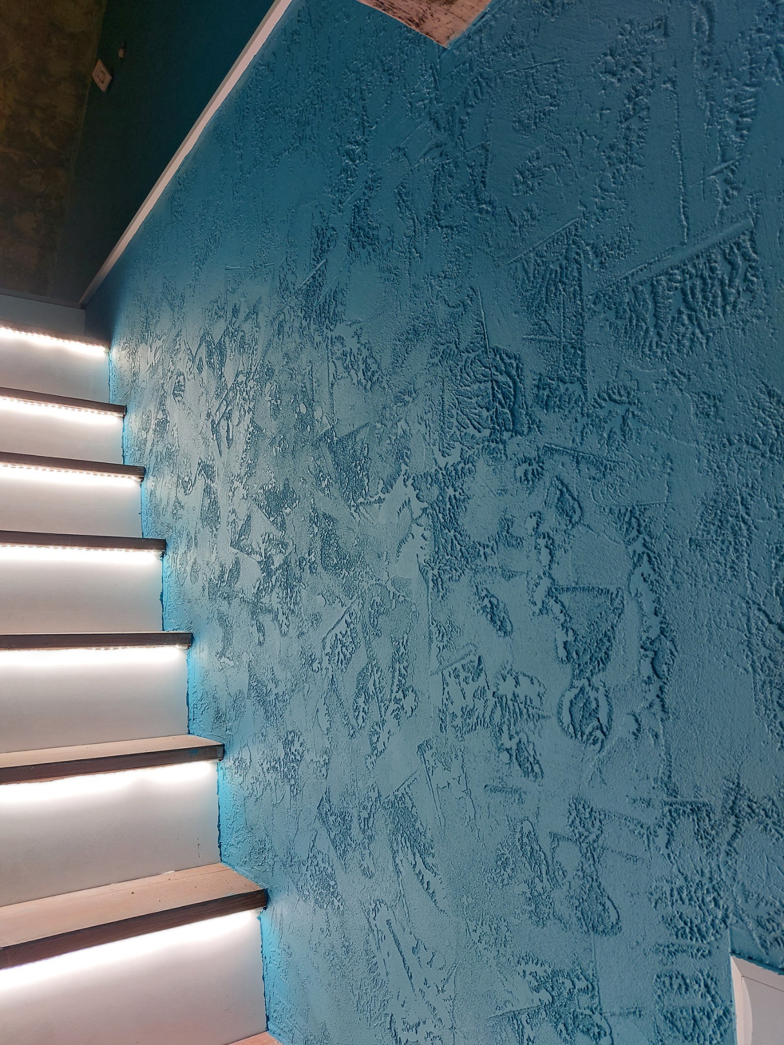 Ściana o nieregularnej fakturze w odcieniach błękitu sąsiadująca z podświetlonymi schodami.